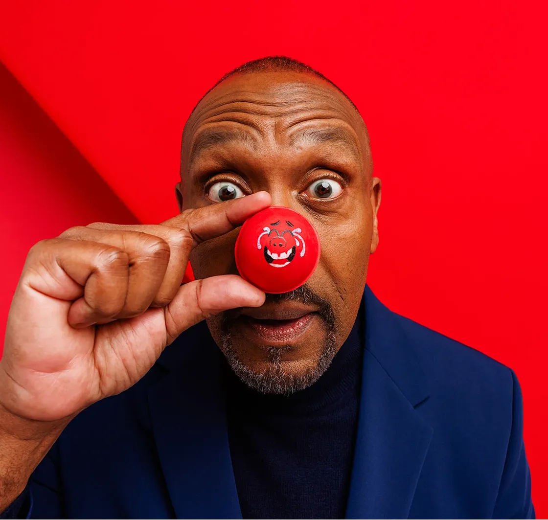 Man in blue jacket holding a red clown nose with a funny face illustration close to his nose against a red background.
