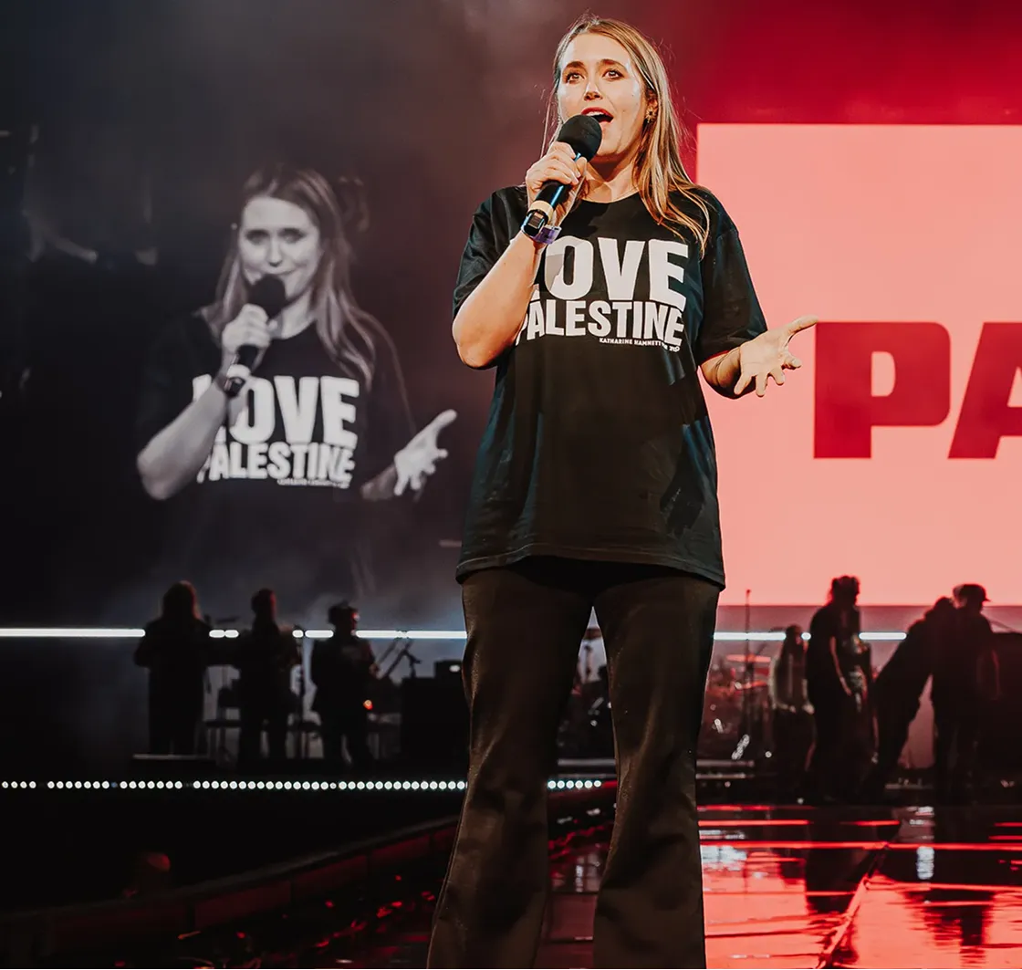 A woman speaking on stage with a microphone wearing a black T-shirt that says 'LOVE PALESTINE' during a concert or event.