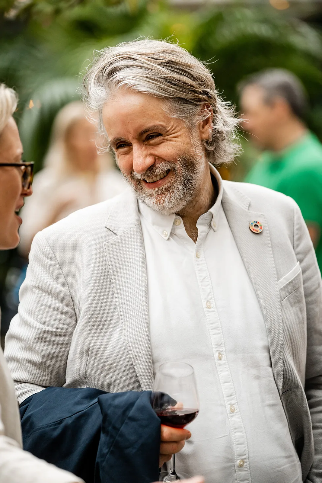 Smiling man with gray hair and beard wearing a light blazer and holding a glass of red wine at a social gathering.
