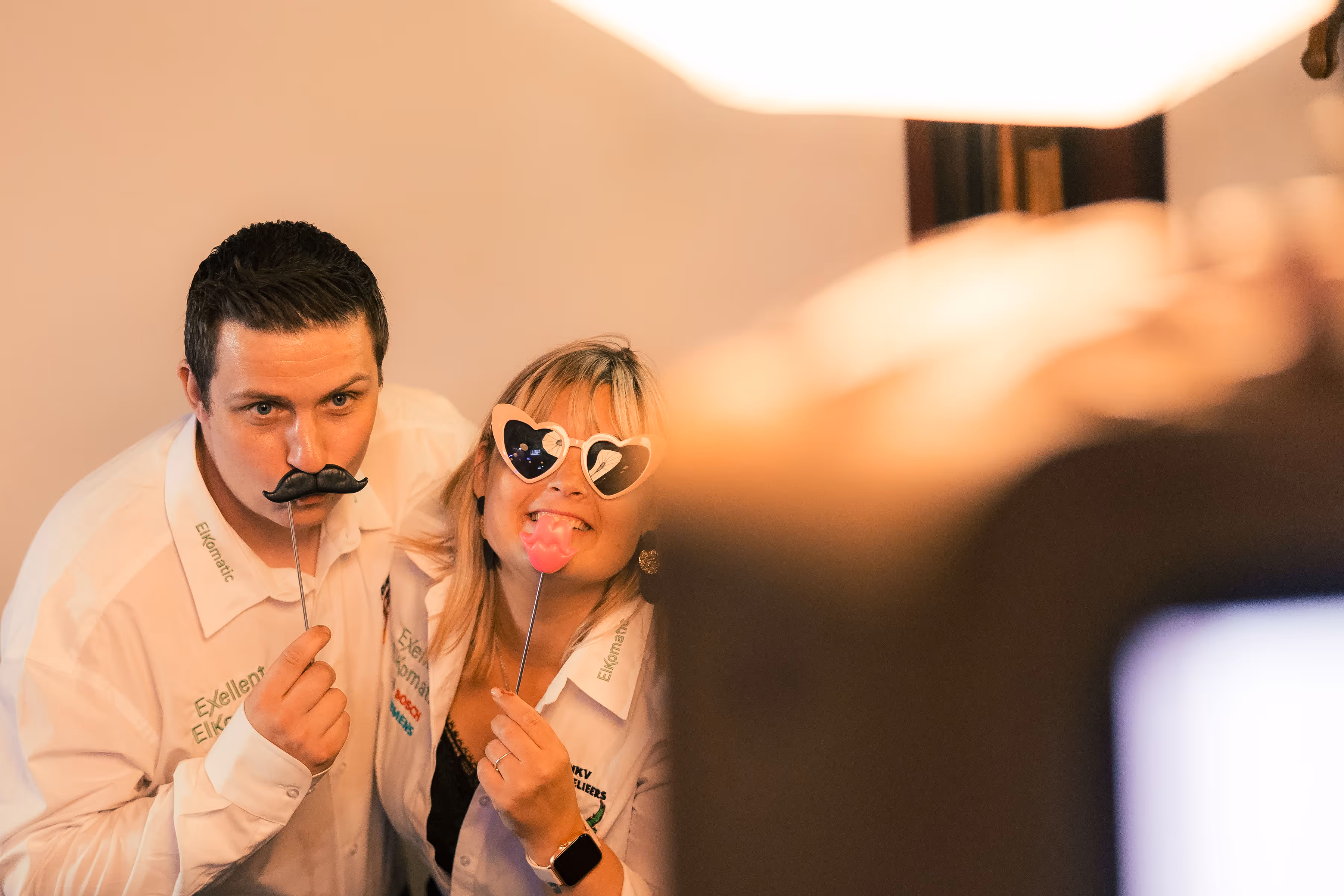 Twee mensen poseren in een photobooth met speelse attributen, waaronder een nepsnor en roze lippen op stok, dragen bedrijfsshirts met logo's.