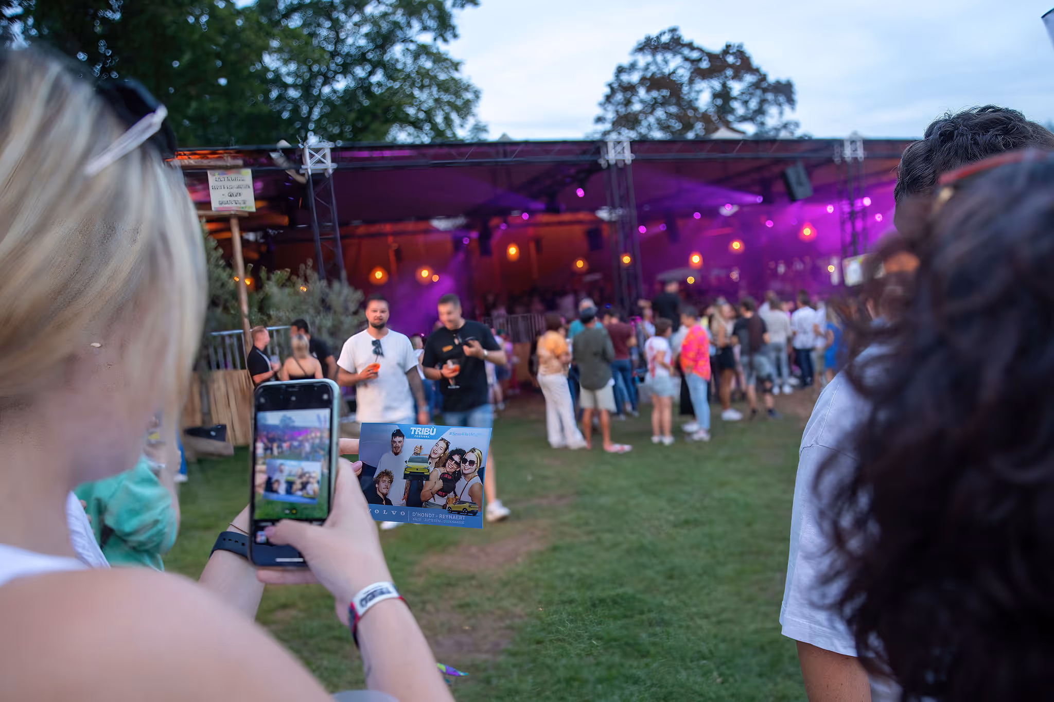 Een vrouw maakt een foto met haar smartphone van een flyer bij een buitenconcert met mensen die ontspannen en praten bij een verlichte tent.