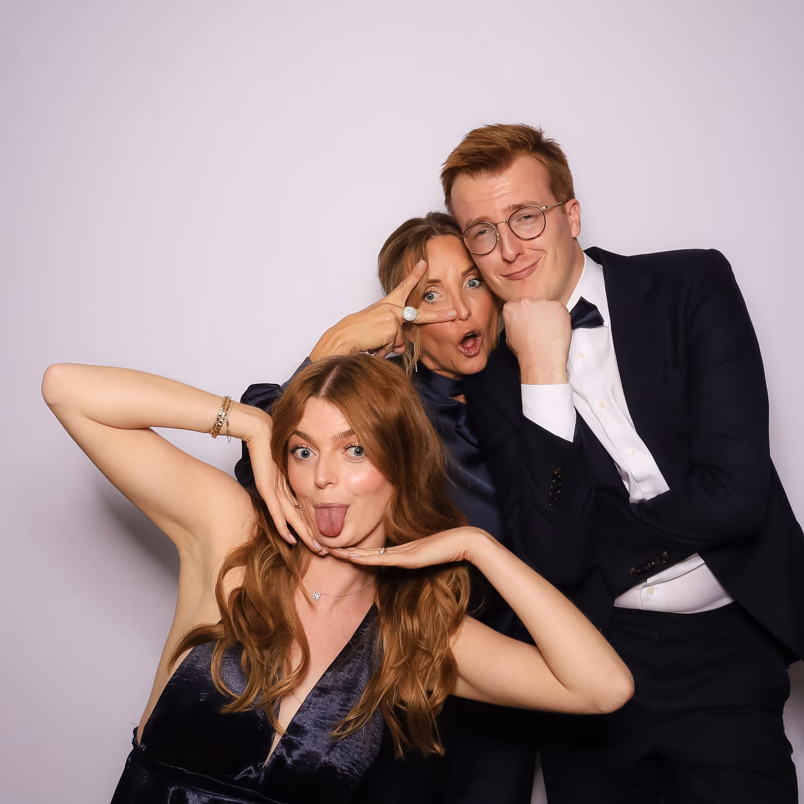 Drie mensen in avondkleding poseren speels voor een witte achtergrond, één vrouw steekt haar tong uit, een andere maakt een V-teken bij haar oog, en een man leunt met zijn kin op zijn vuist.