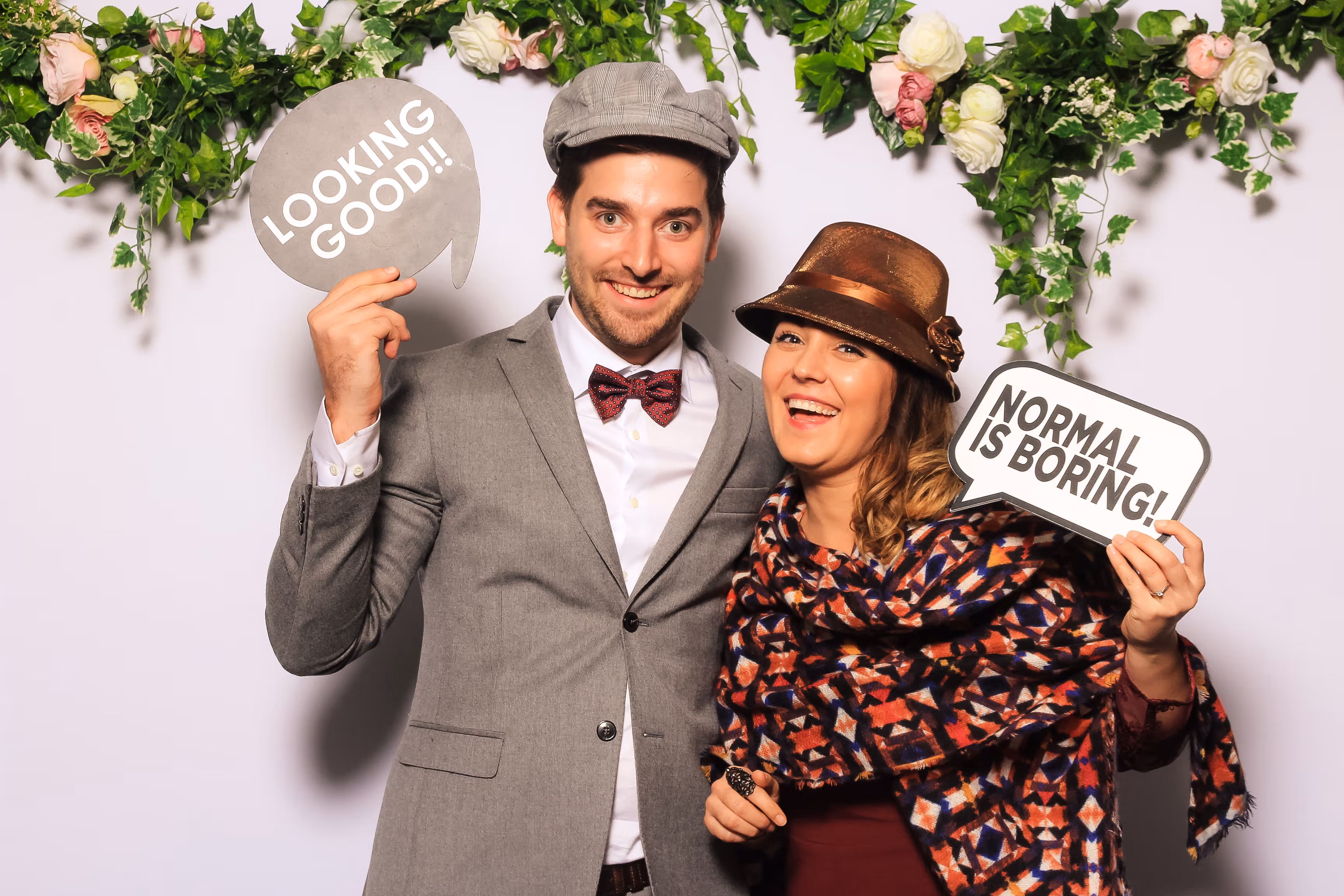 Een man en vrouw poseren lachend met hoeden en tekstborden in een fotohokje met bloemen decor.