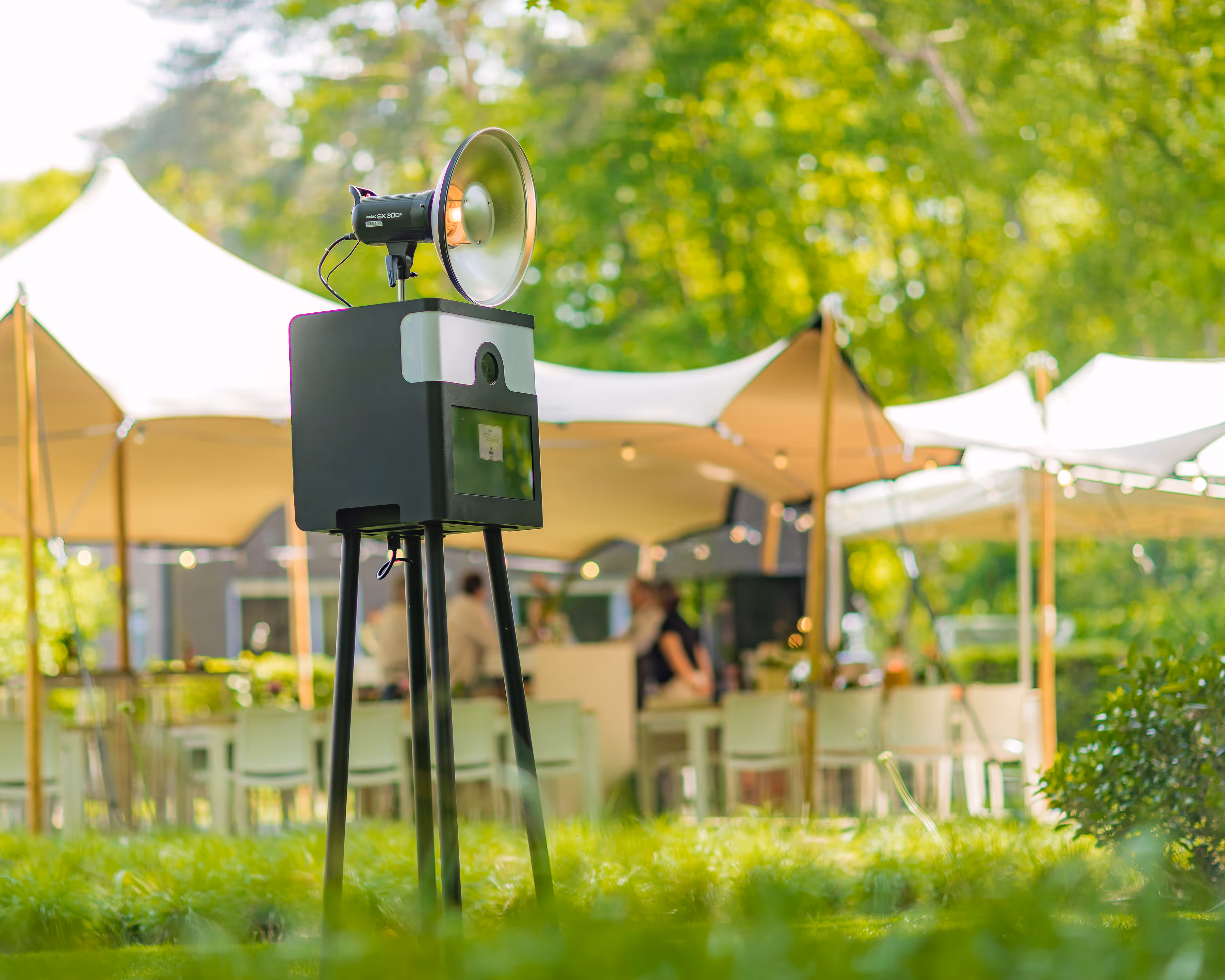 Fotohokje met flitserstaander buiten in een tuin bij een evenement met witte tenten en mensen op de achtergrond.