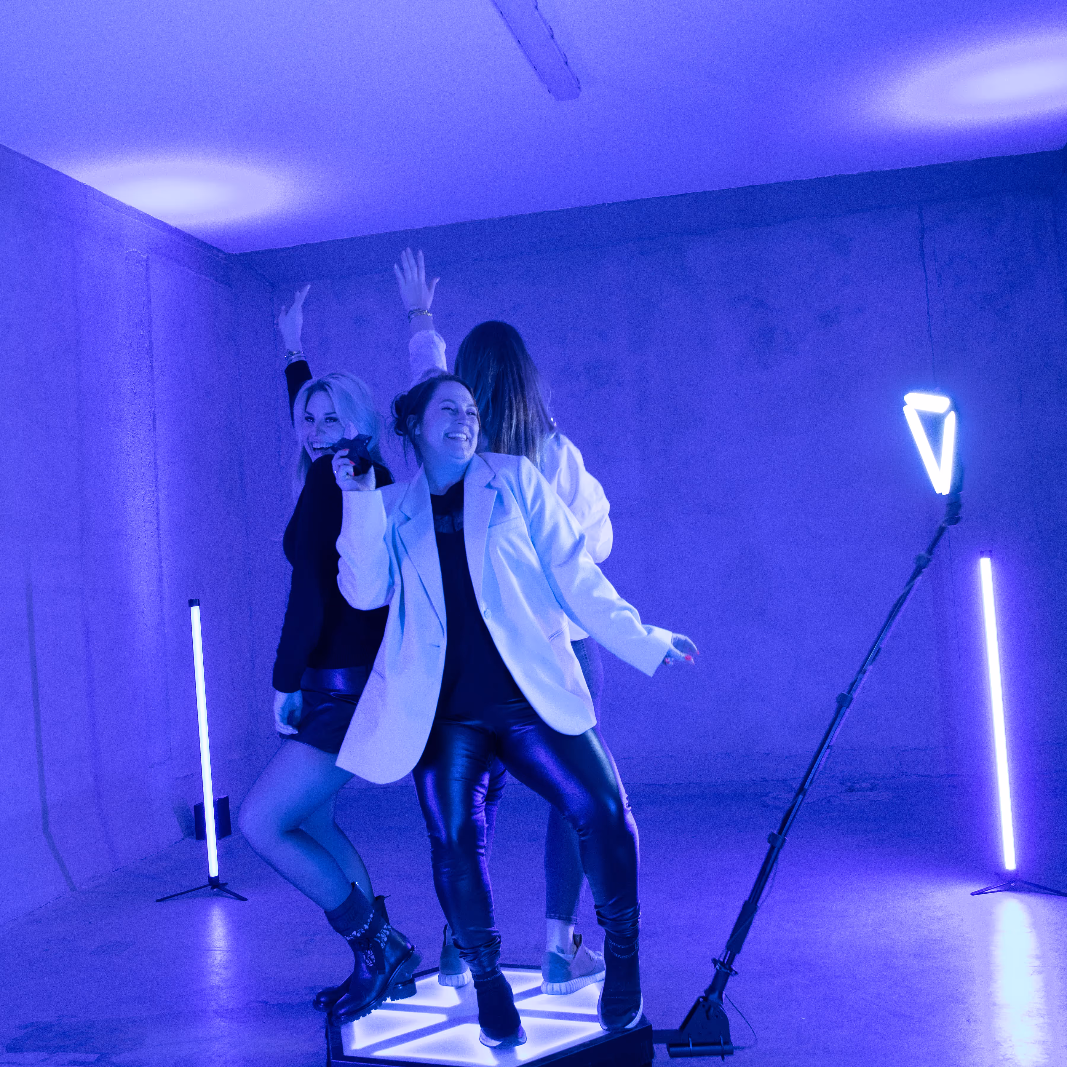 Drie jonge vrouwen dansen en lachen op een verlicht platform in een kamer met paarse neonverlichting.
