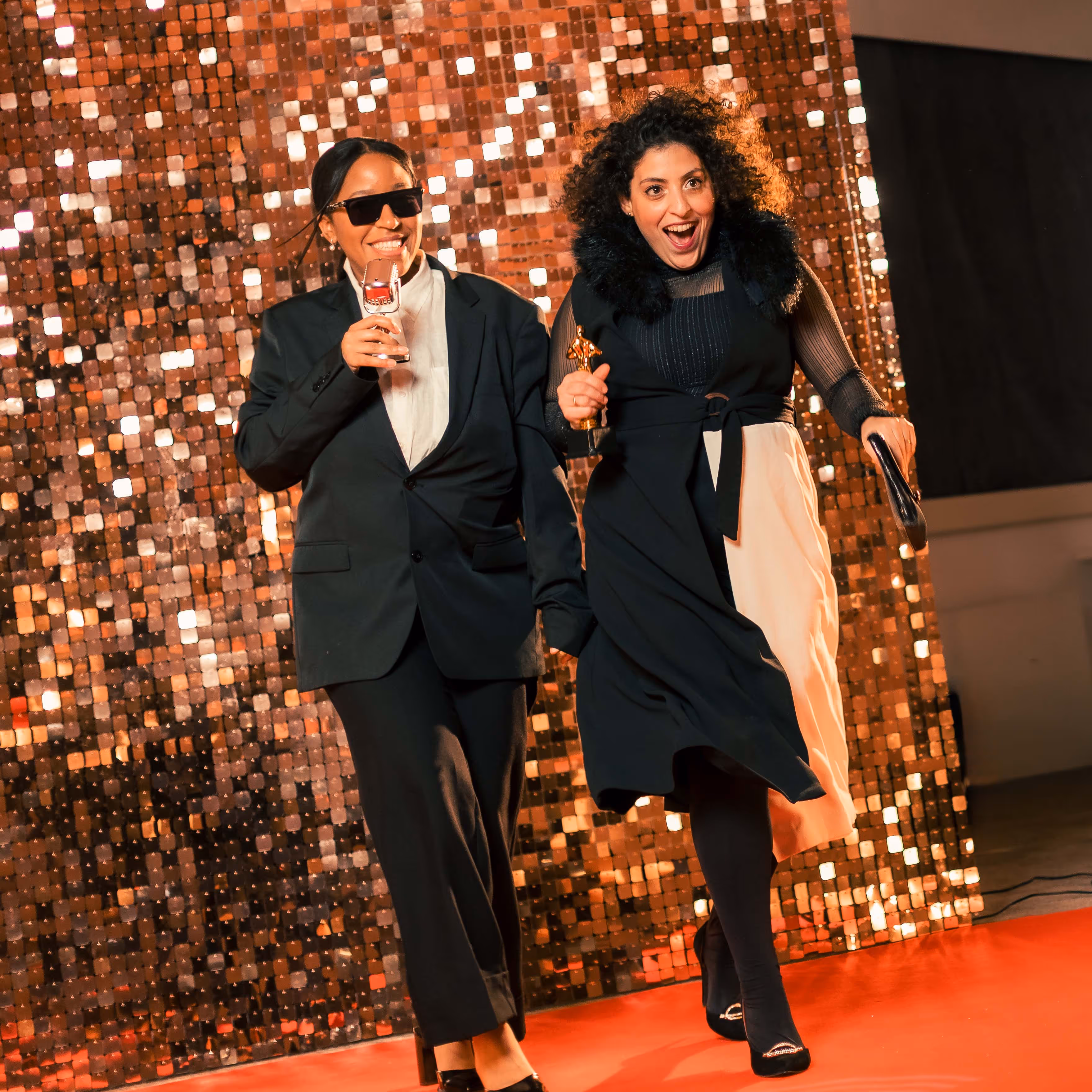Twee feestelijk geklede vrouwen lopen over een rode loper, één met een glas champagne en de ander met een prijsbeeldje en een clutch.