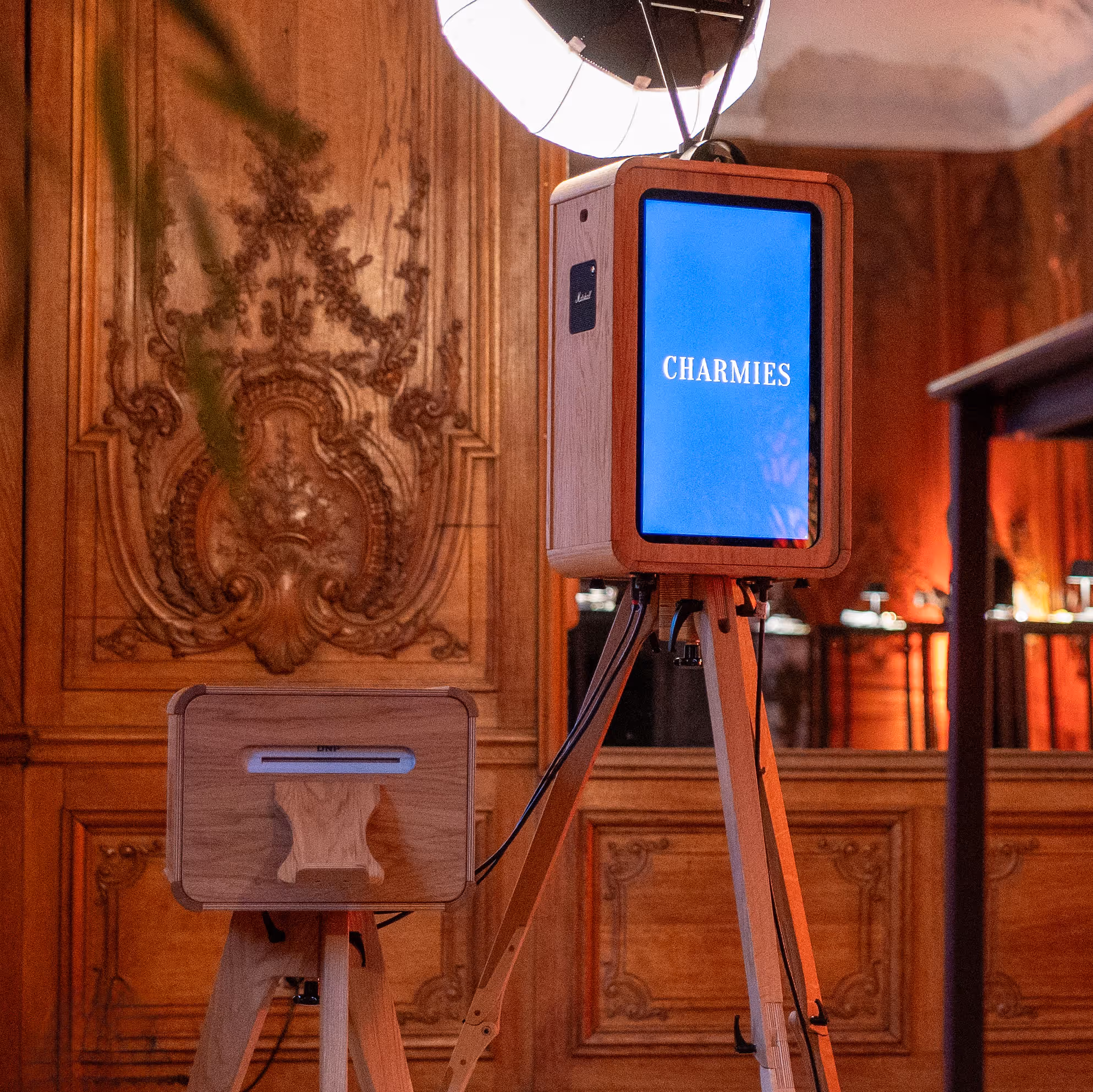 Fotocabine met houten omlijsting en blauw scherm met tekst 'CHARMIES' in een kamer met sierlijke houten panelen.