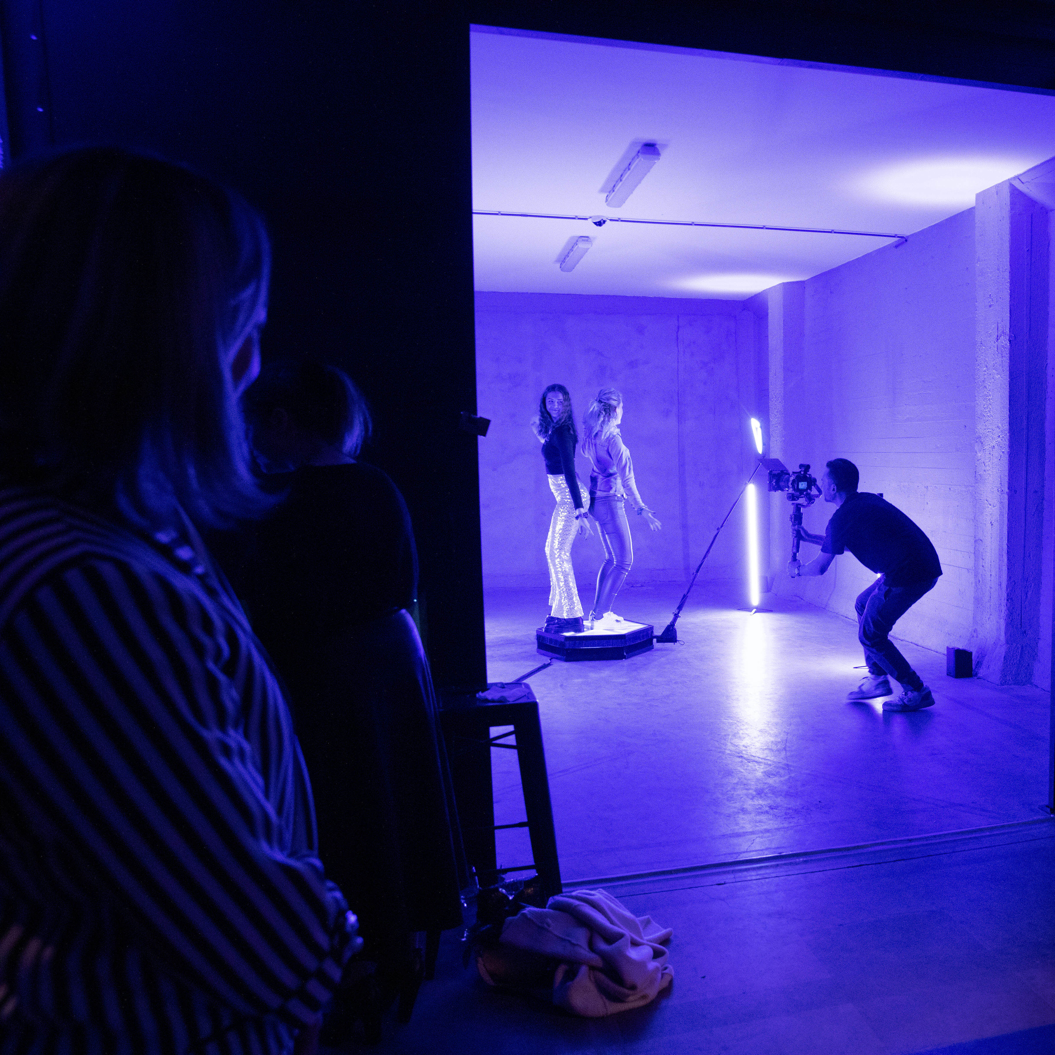 Een fotograaf maakt een foto van twee vrouwen die op een platform staan in een paarse verlichte studio, terwijl twee mensen toekijken in de schaduw.