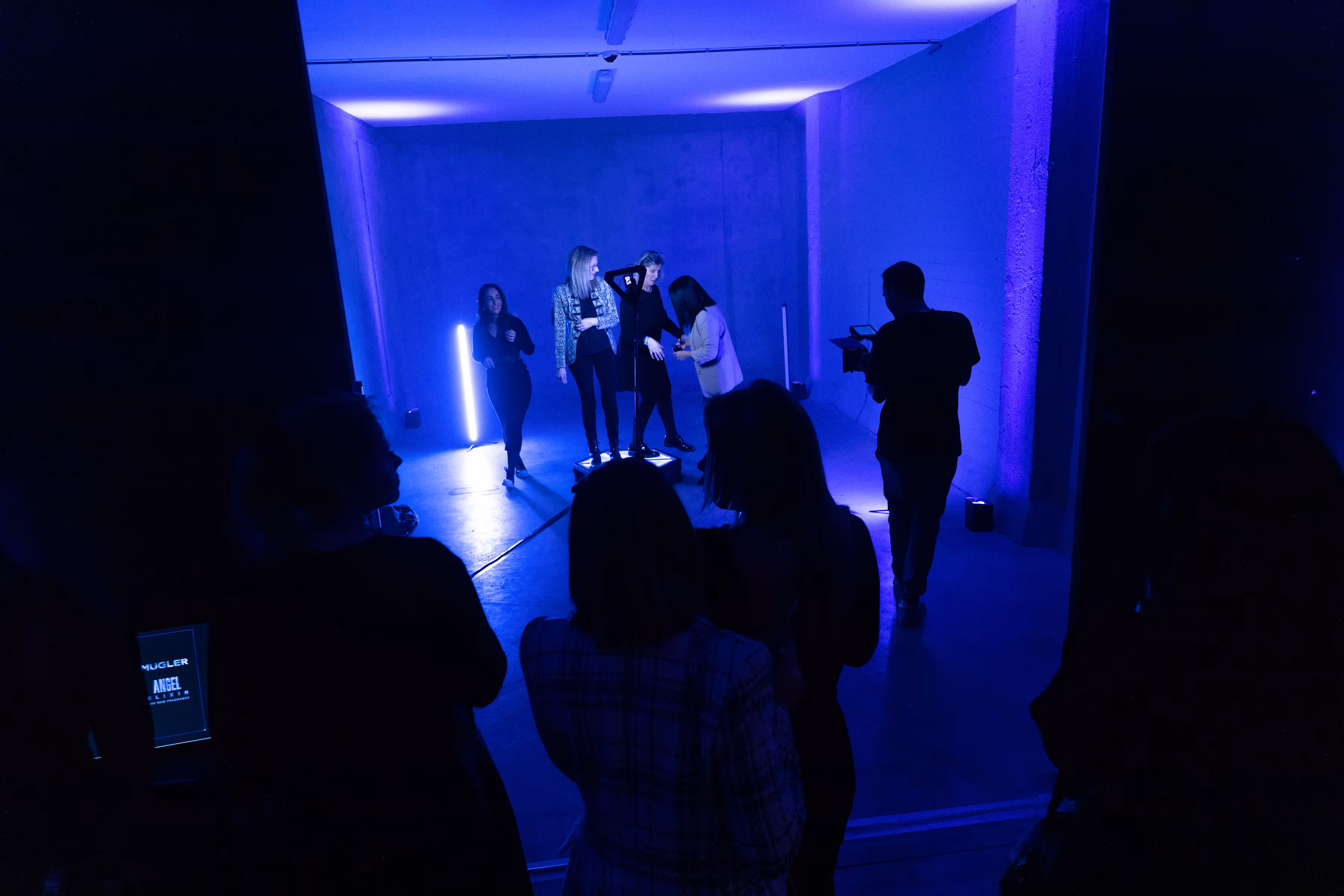 Mensen in een donkere kamer met blauwe verlichting, sommige poseren terwijl anderen filmen of toekijken.