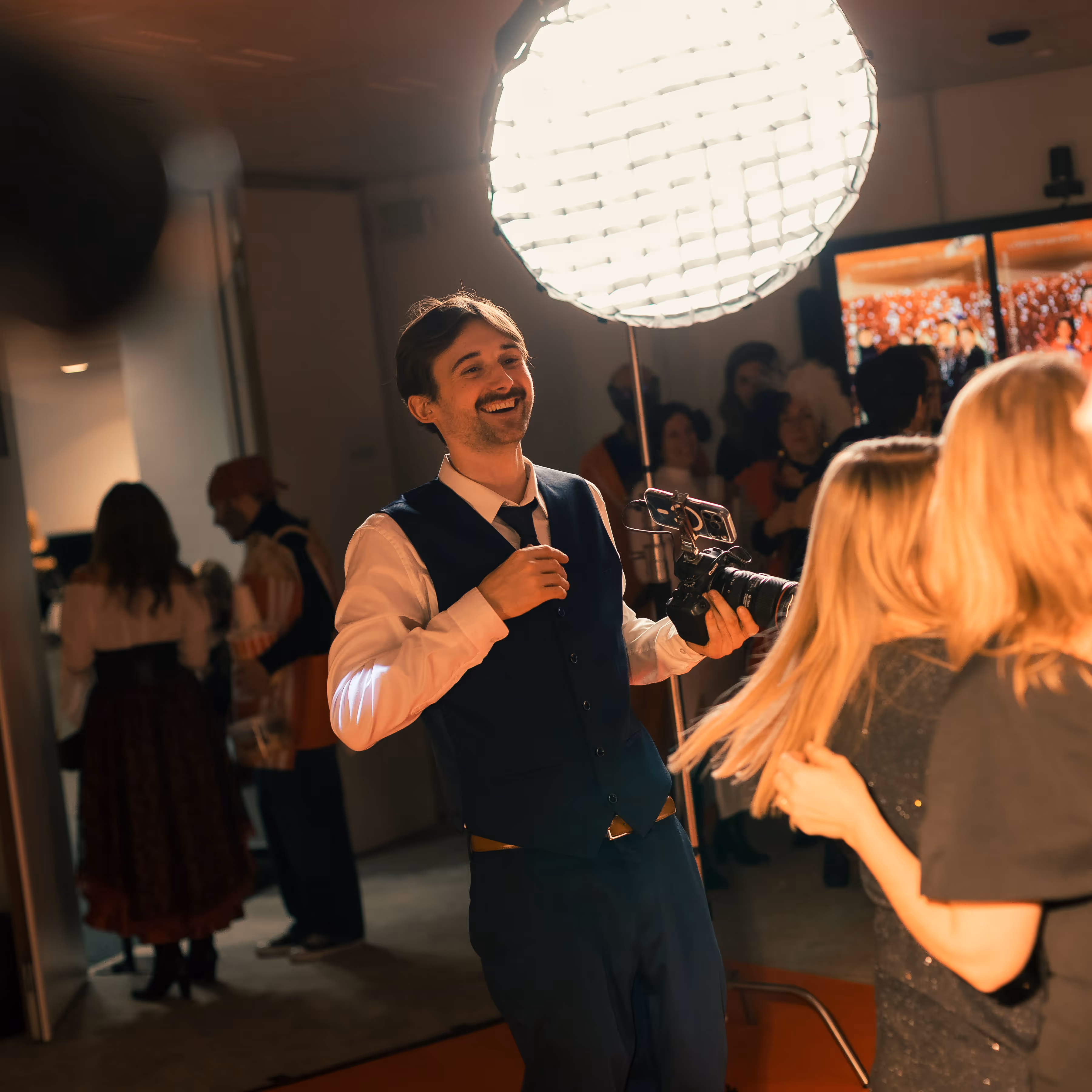Vrolijke man in een donkerblauw pak filmt een evenement met een camera onder een grote fotolicht, omringd door feestende mensen.