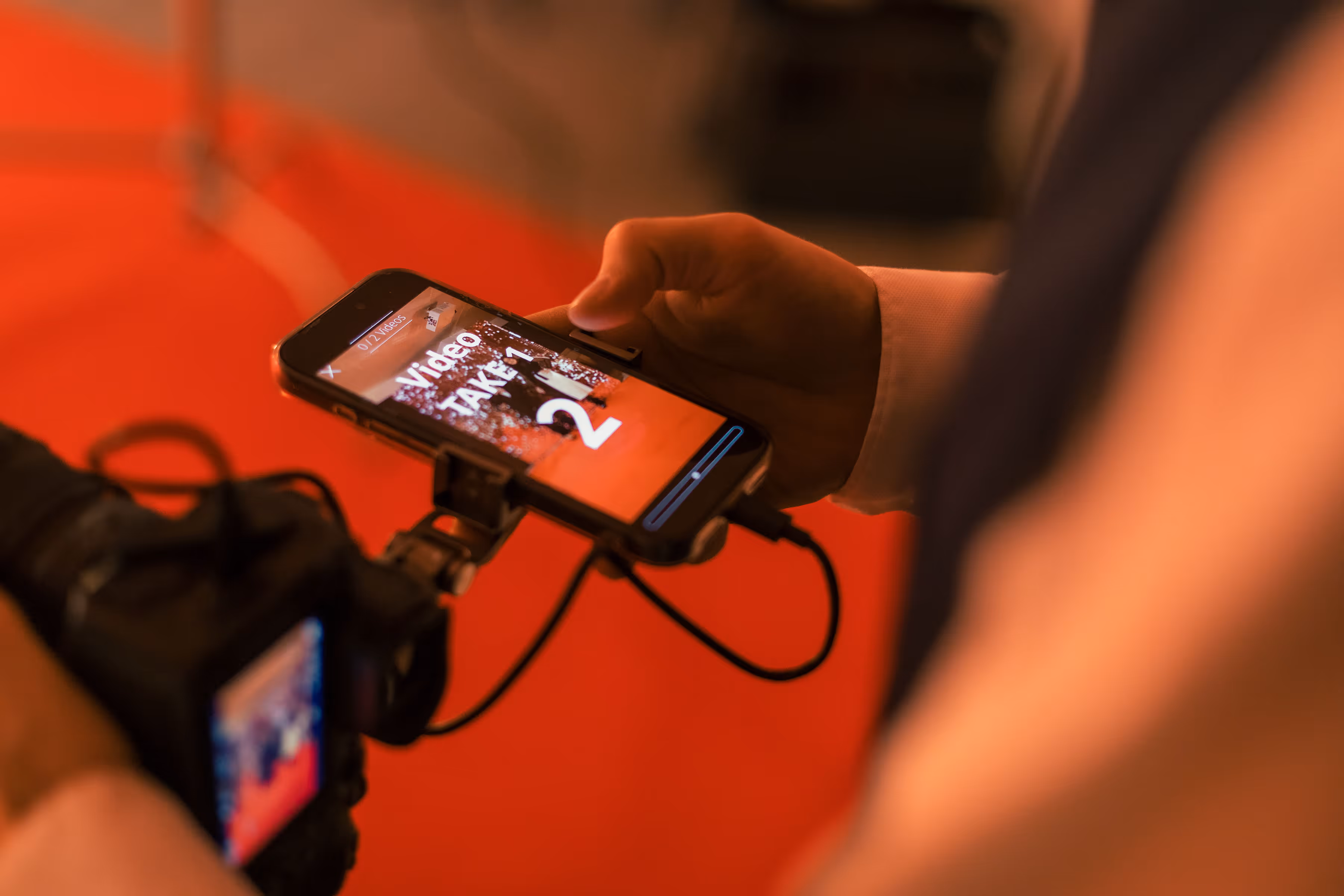 Hand van een persoon die een smartphone vasthoudt met een videospeler die 'Video TAKE 1 2' toont tegen een rode achtergrond.
