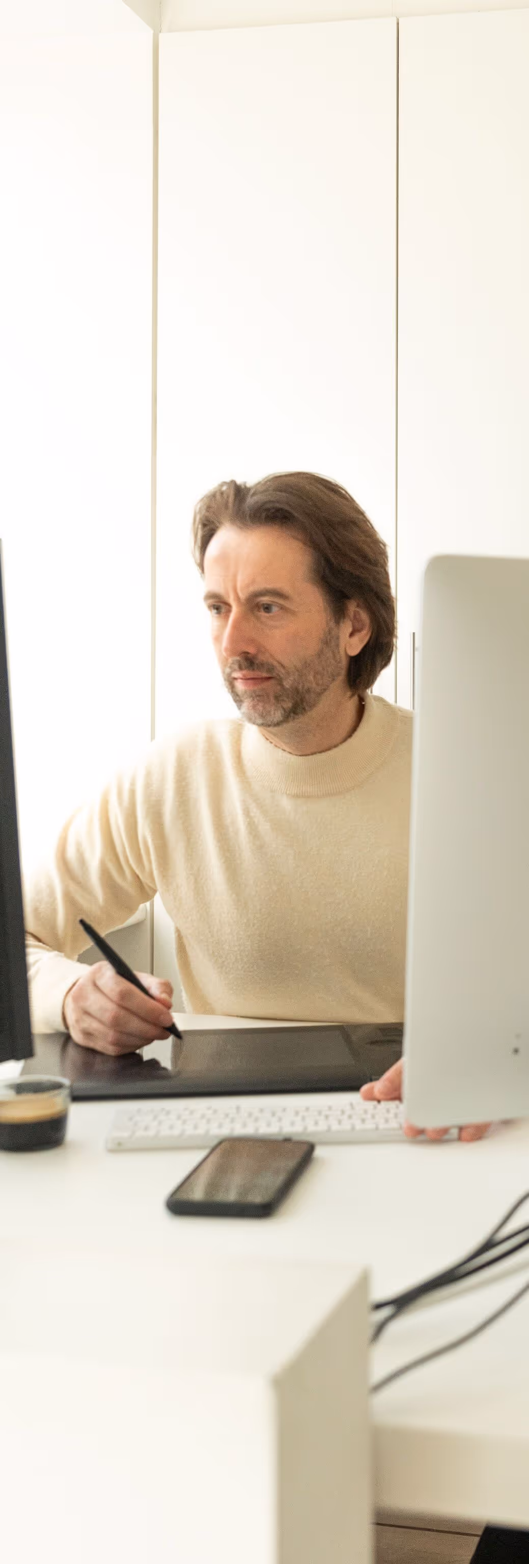 Man in beige sweater using a digital drawing tablet at a desk with two monitors and a smartphone.