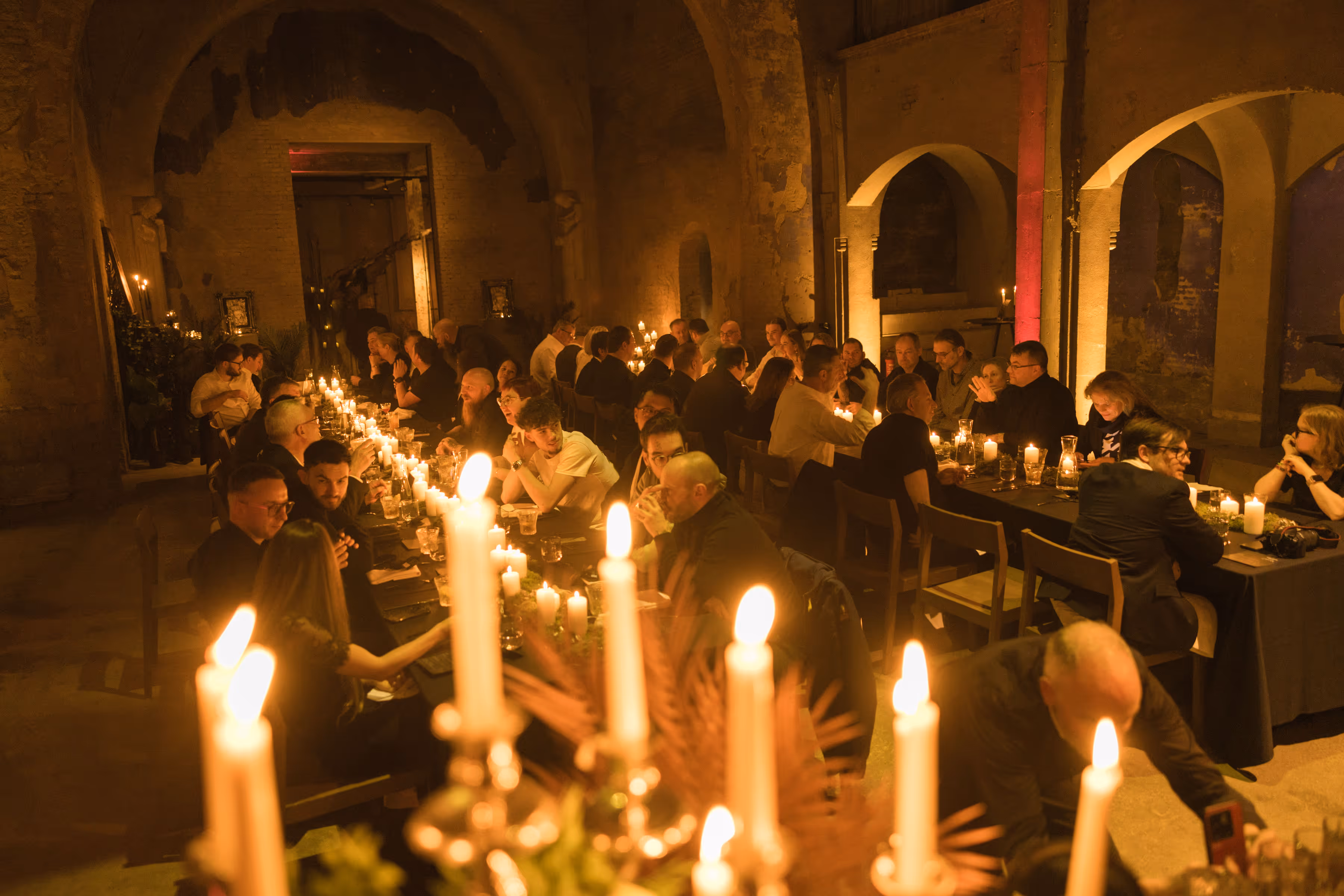 Gezelschap zit aan lange tafels met vele brandende kaarsen in een sfeervol verlichte, oude stenen zaal.