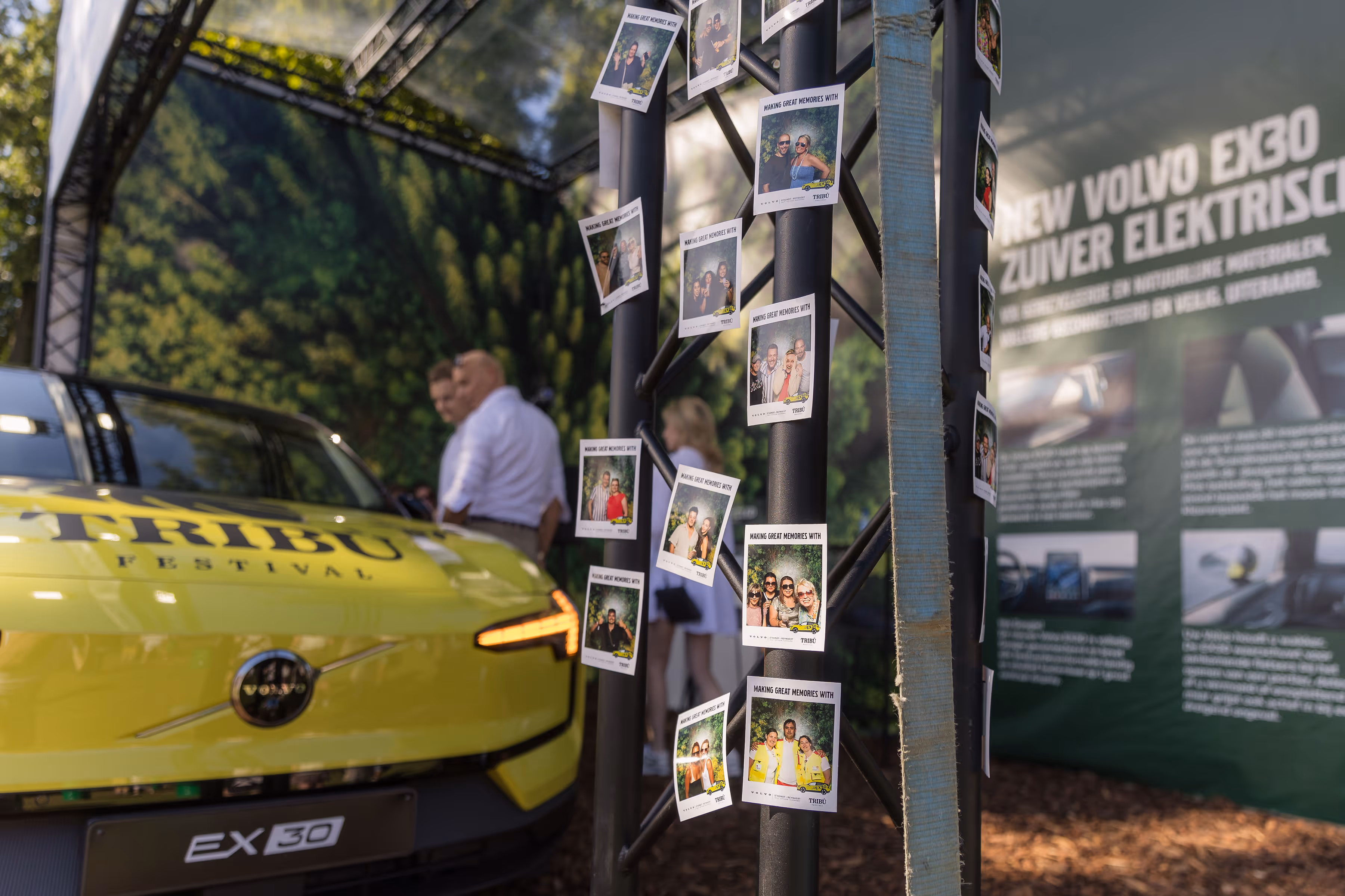 Felgeleide gele Volvo EX30 elektrisch voertuig naast paal met foto’s van festivalbezoekers en banner met details over de nieuwe Volvo EX30.