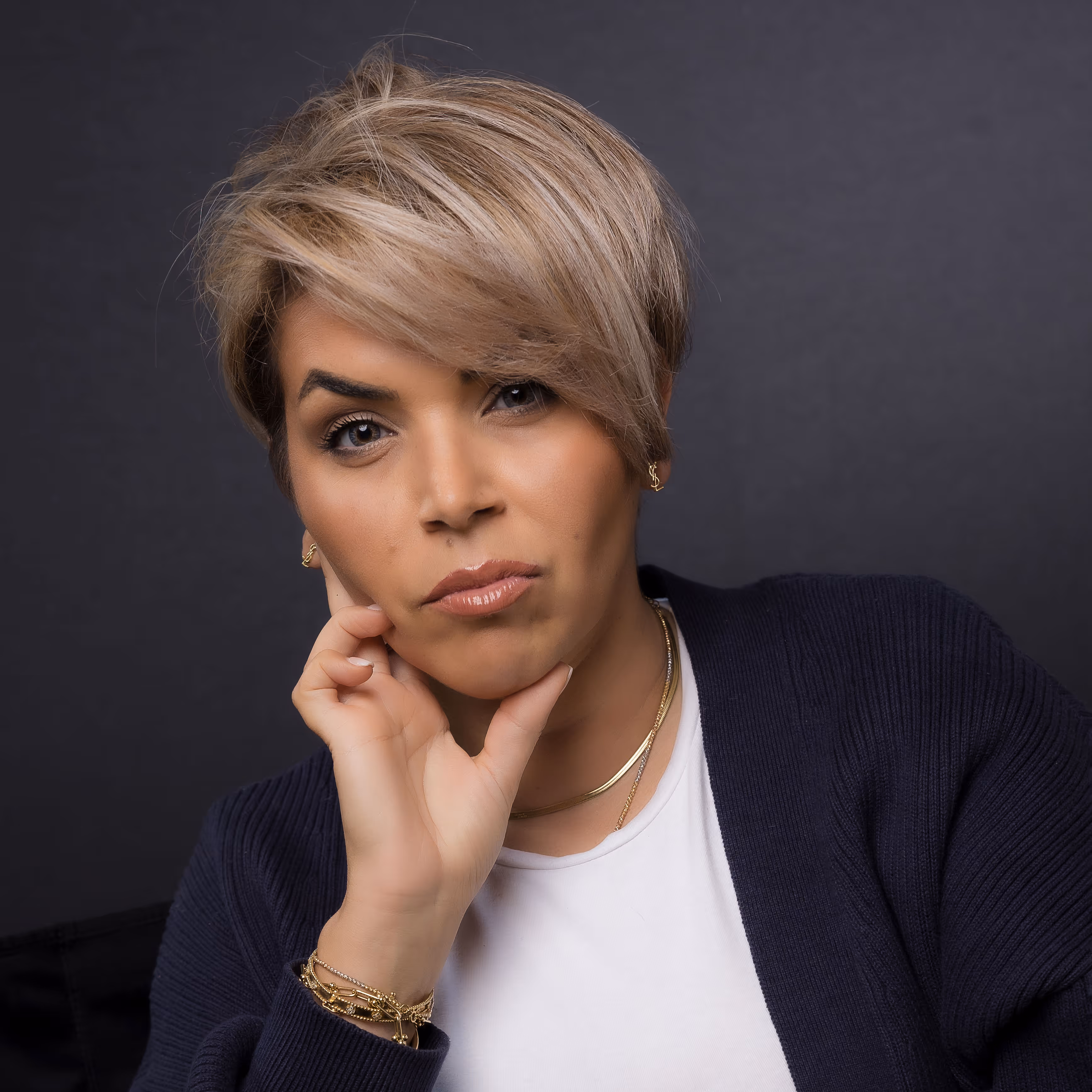 Portret van een vrouw met kort blond haar, lichte make-up, een wit T-shirt, een donkerblauw vest en gouden sieraden, tegen een donkere achtergrond.