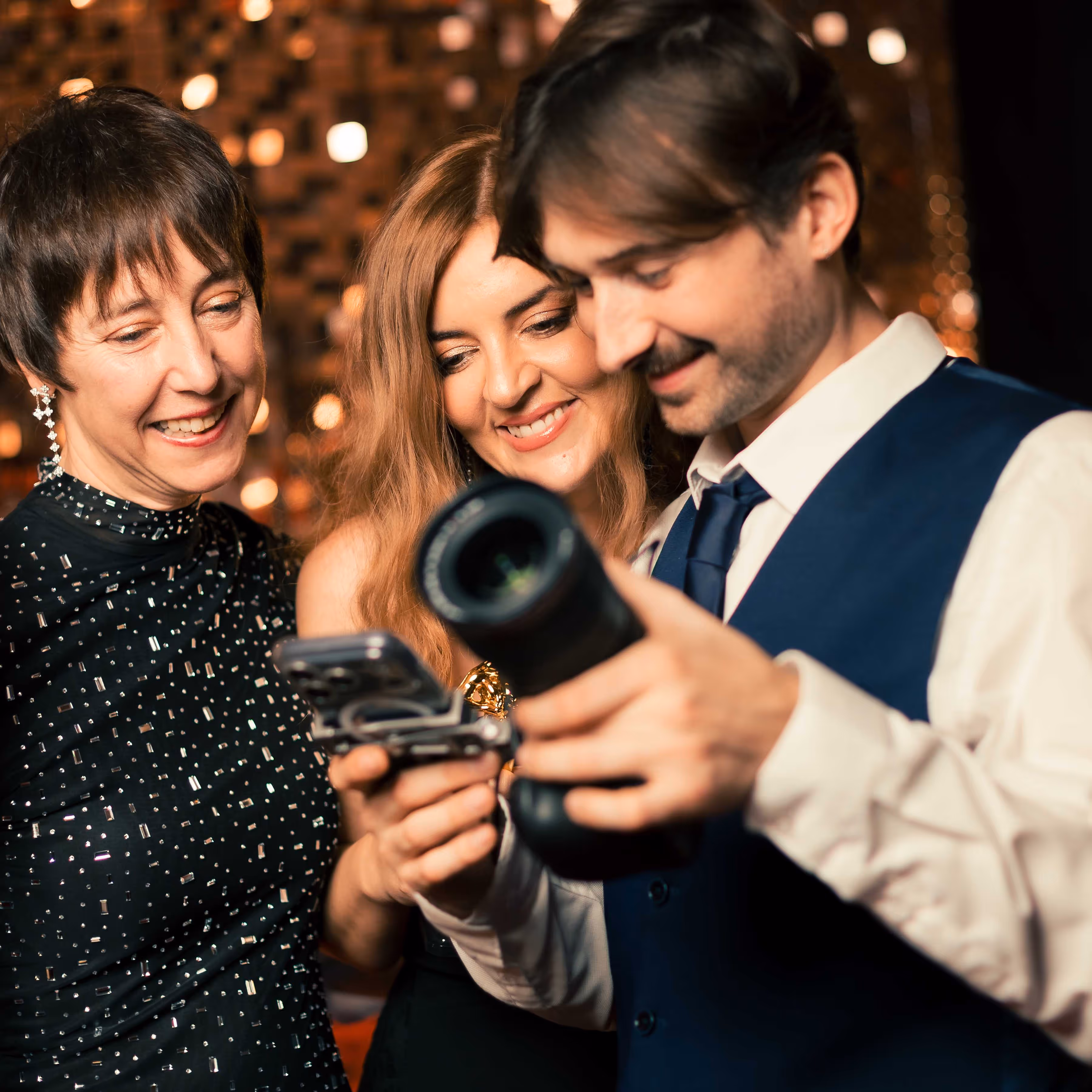 Drie feestelijk geklede volwassenen bekijken samen een camera en een smartphone met een warme achtergrondverlichting.