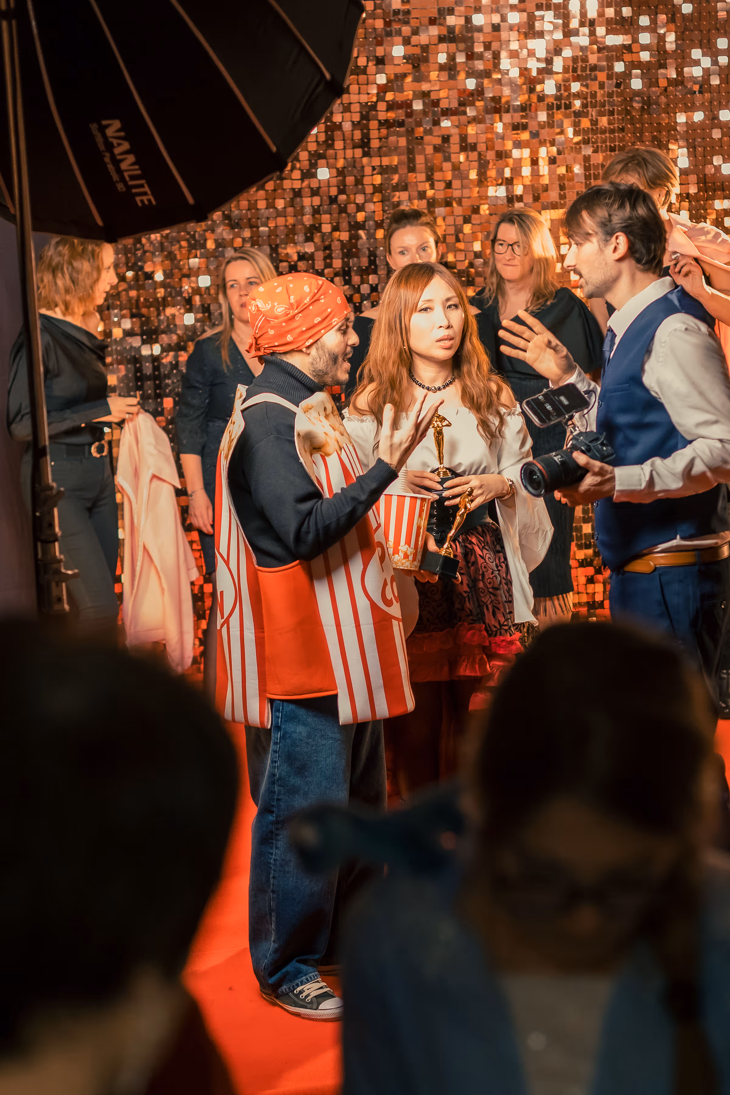 Mensen in een fotoshoot op een rode loper met een man verkleed als popcorn en een vrouw die een filmprijs vasthoudt.