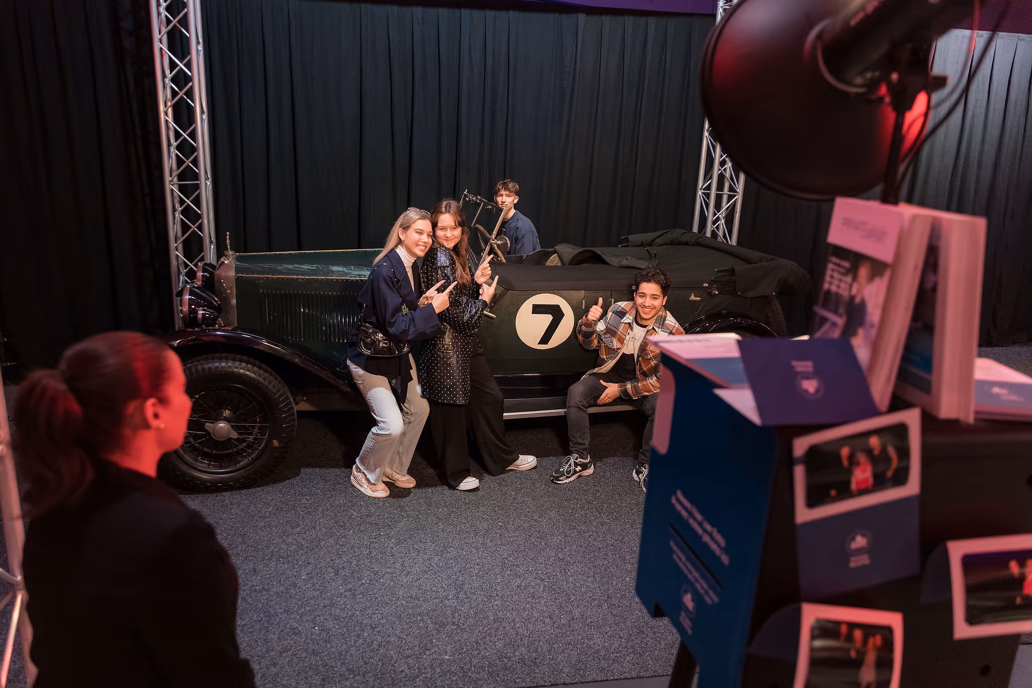 Vier jonge mensen poseren lachend bij een klassieke auto met het nummer 7 op de deur in een binnenruimte met zwarte gordijnen.
