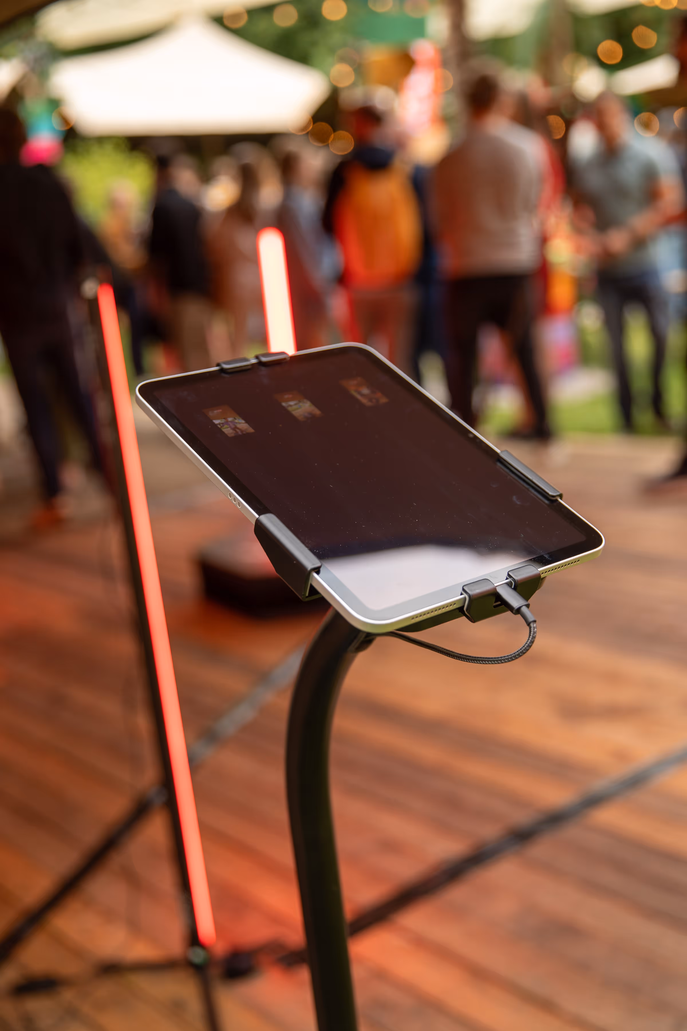 Tablet op standaard met aanhangende kabel, op een houten vloer met een groep mensen op de achtergrond buiten.