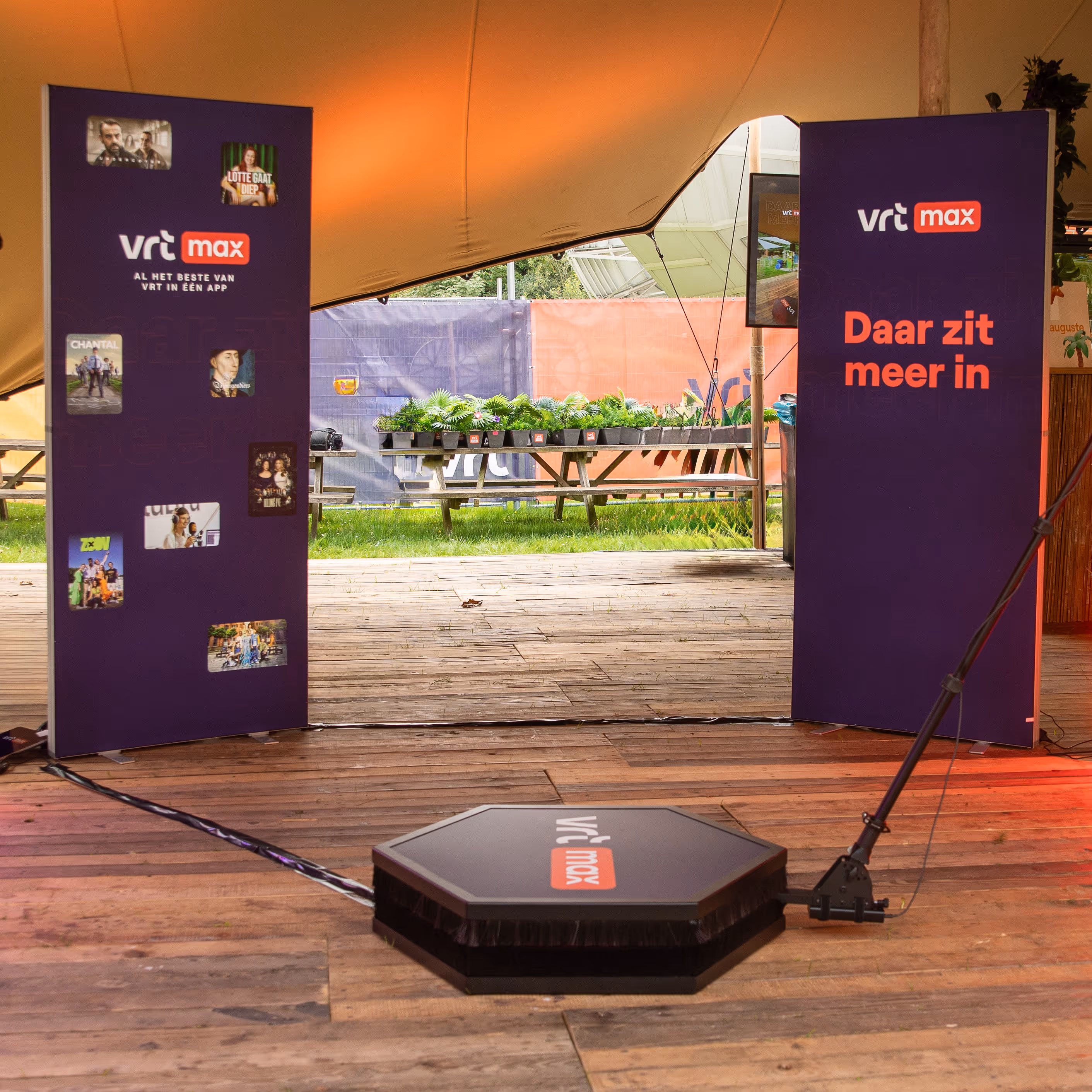Twee paarse banners met VRT Max logo en reclame, geplaatst onder een tent met zicht op houten vloer en buiten een picknicktafel met planten.