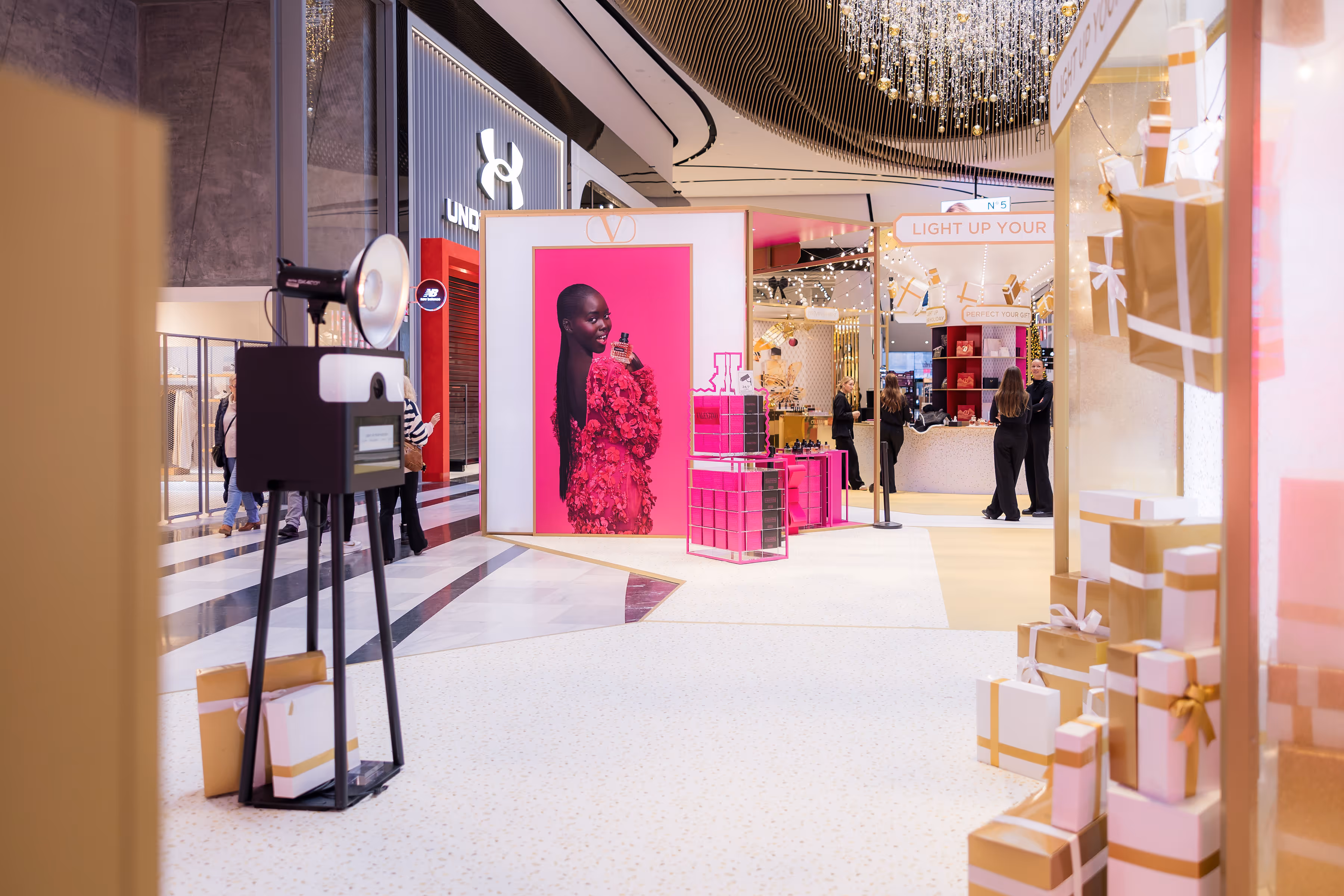 Interieur van een winkelcentrum met een fotohokje, een groot roze reclamebord met een vrouw in een roze jurk, en diverse ingepakte cadeaus.