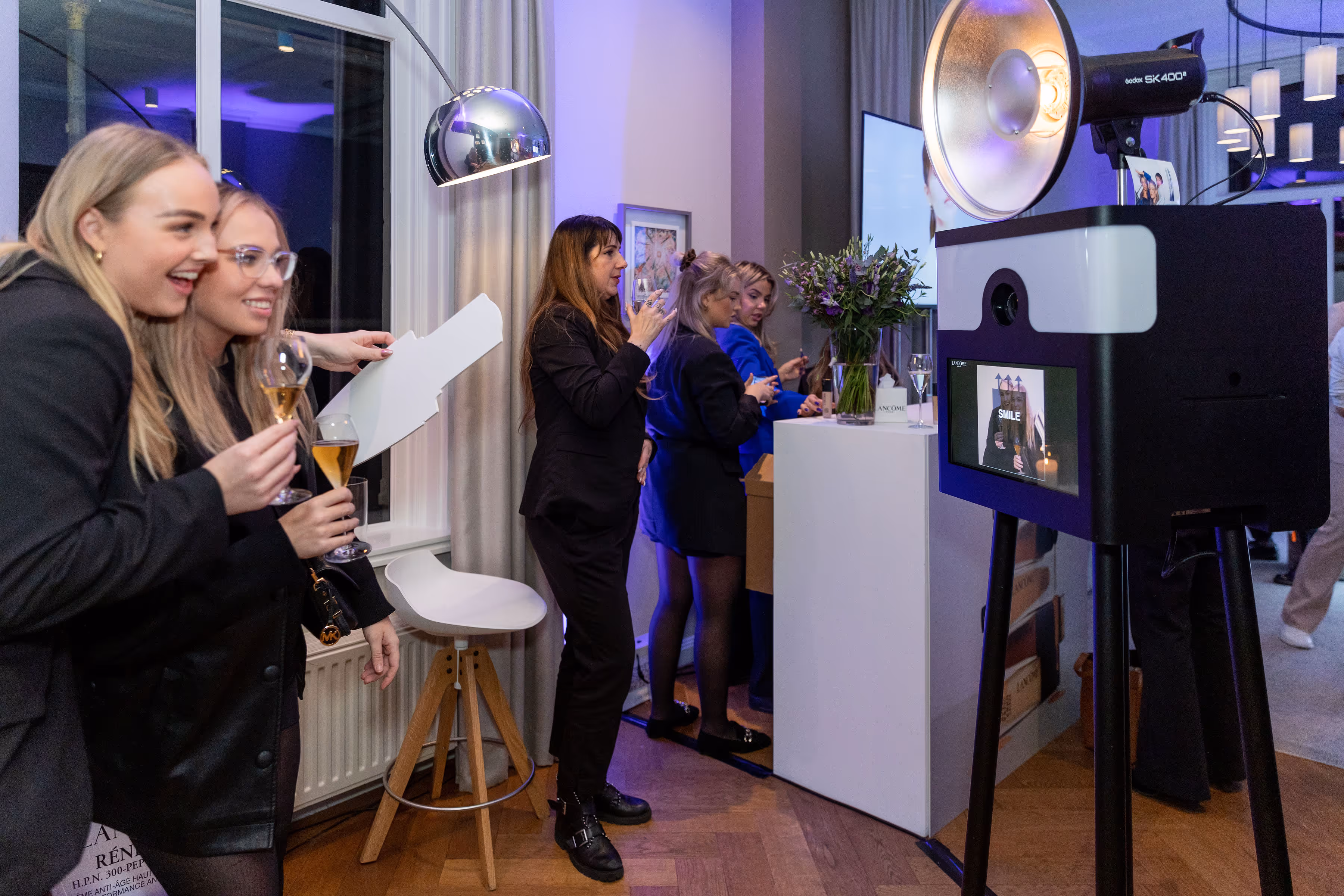 Groep vrouwen geniet van een drankje en lachen bij een fotohokje tijdens een avondfeest.