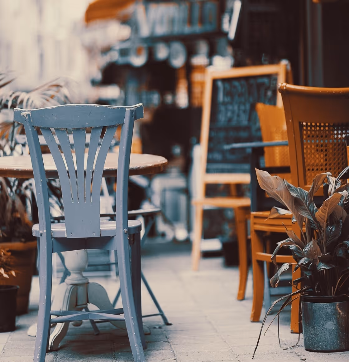 Außensitzbereich eines Cafes mit grauem Holzstuhl, Tisch, weiteren Stühlen und einem Topfpflanze.