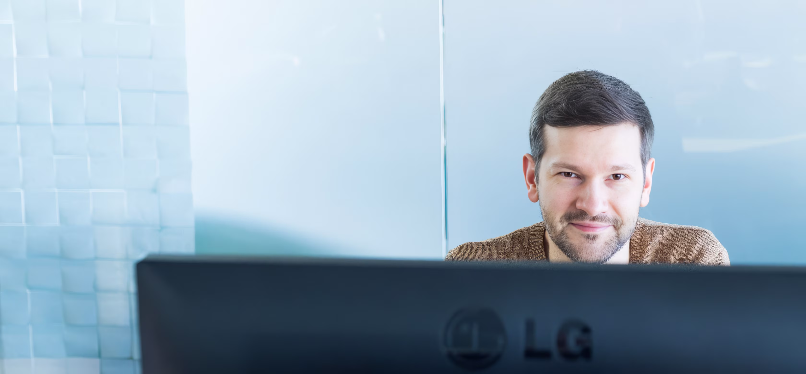 Mann mit braunem Pullover sitzt lächelnd hinter einem LG-Computerbildschirm an einem hellen Arbeitsplatz.