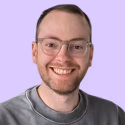 Lächelnder Mann mit Brille und grauem Pullover vor lavendelfarbenem Hintergrund.