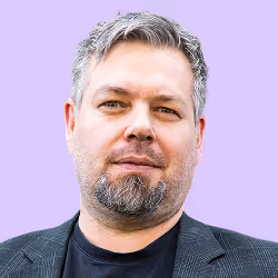 Porträt eines Mannes mit kurzgeschnittenem grauem Haar und Bart vor lila Hintergrund.