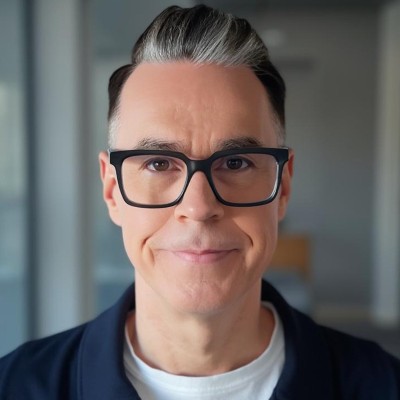 Porträt eines Mannes mit schwarzen Brillen, grauem Haaransatz und leichtem Lächeln vor verschwommenem Hintergrund.