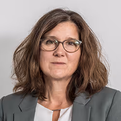 Frau mit braunen, lockigen Haaren, Brille und grauem Blazer vor grauem Hintergrund.
