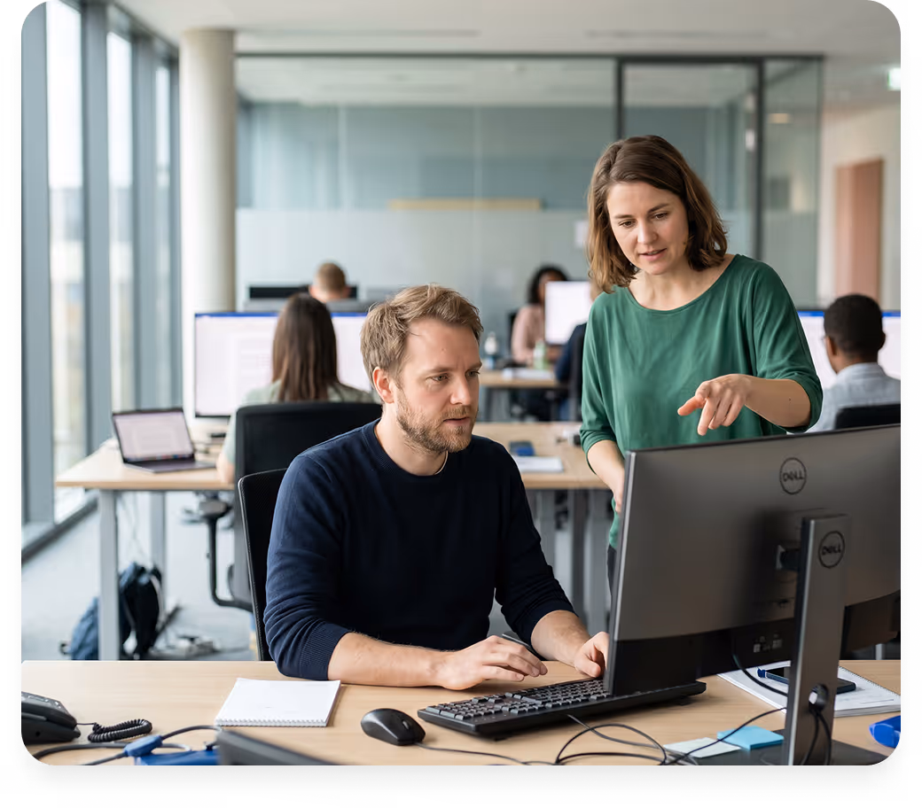 Eine Frau erklärt einem Mann etwas am Computer in einem modernen Büro mit mehreren Mitarbeitern.