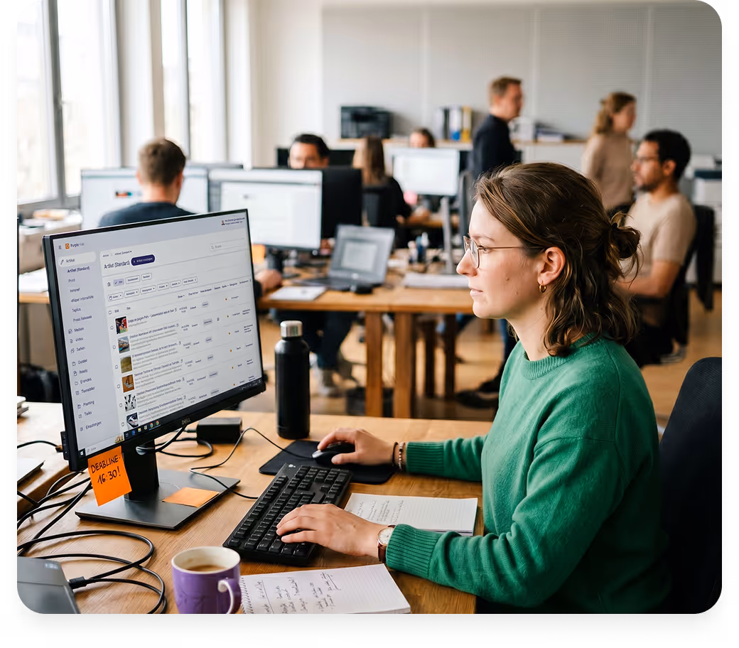 Frau in grünem Pullover arbeitet konzentriert am Computer in offenem Büro mit Kollegen im Hintergrund.