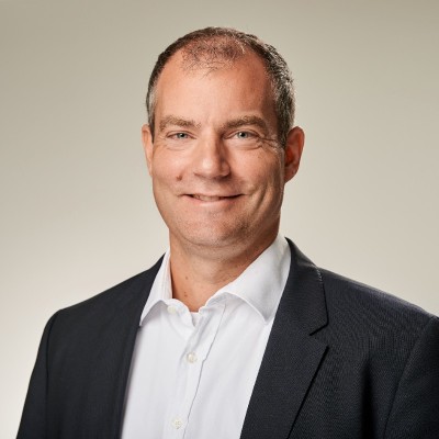 Porträt eines lächelnden Mannes in weißem Hemd und schwarzem Sakko vor neutralem Hintergrund.