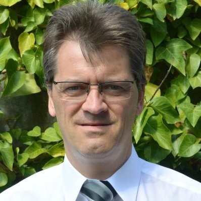 Porträt eines Mannes mit Brille in weißem Hemd und gestreifter Krawatte vor grünen Blättern.