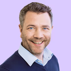 Lächelnder Mann mit kurzen, lockigen braunen Haaren vor hellem lila Hintergrund.