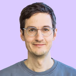 Porträt eines Mannes mit braunen Haaren, Brille und grauem Pullover vor lila Hintergrund.