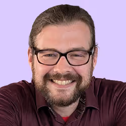 Lächelnder Mann mit Brille, Bart und zurückgebundenen Haaren vor helllila Hintergrund.
