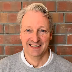 Mann mit hellgrauen Haaren und grauem Pullover lächelt vor Backsteinmauer.
