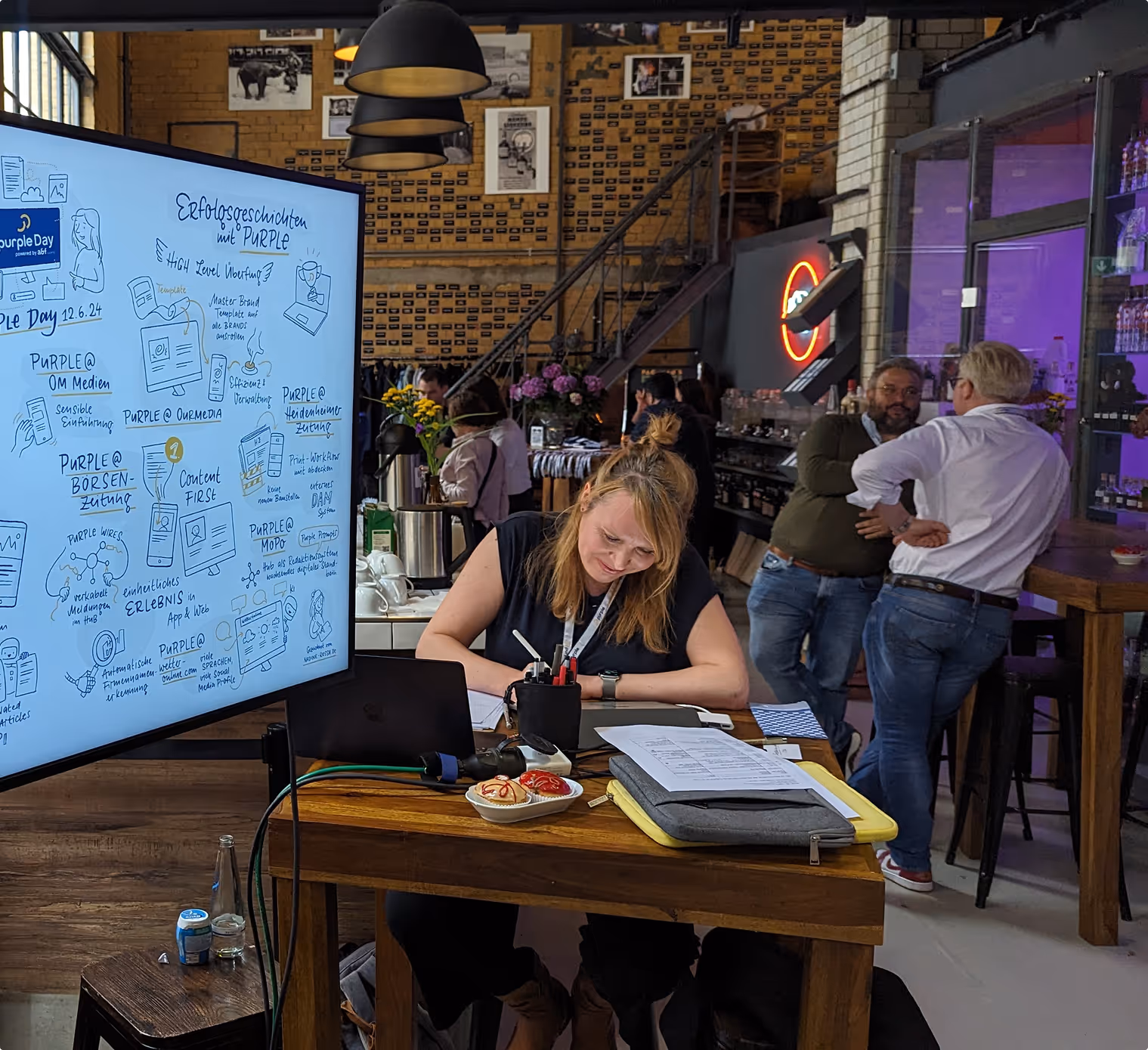 Frau arbeitet an einem Tisch mit Laptop und Notizen, im Hintergrund unterhalten sich Menschen in Café.