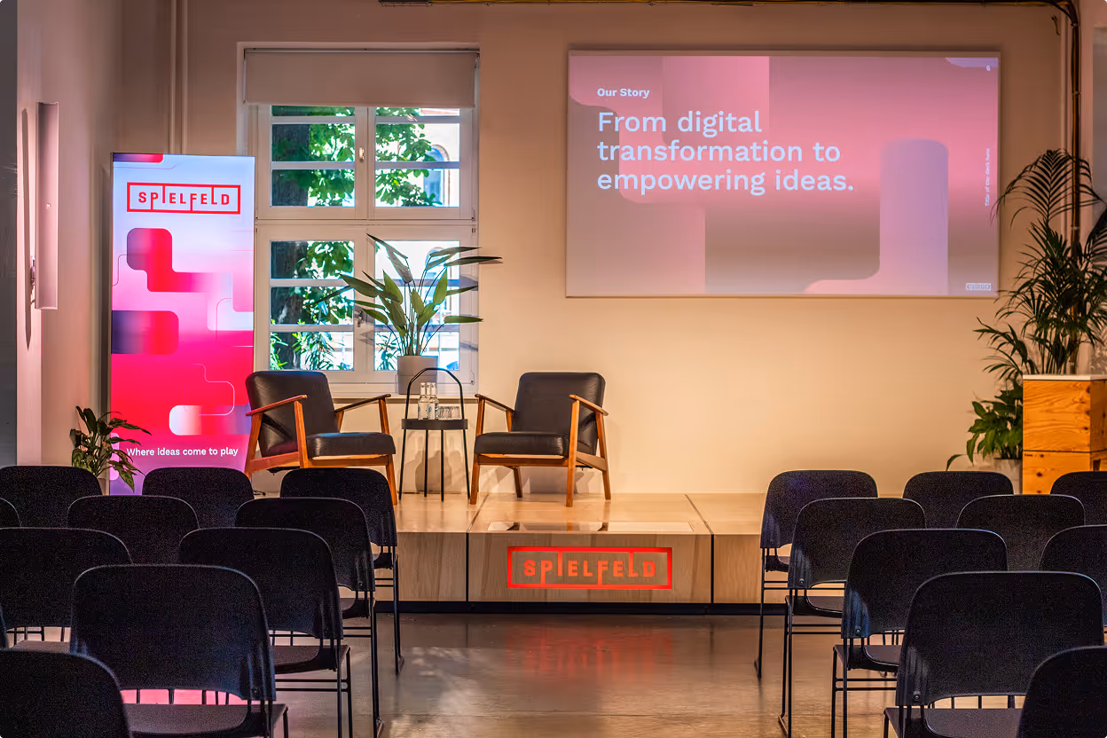 Leerer Konferenzraum mit zwei Sesseln auf Bühne, Bildschirm mit Text zur digitalen Transformation.