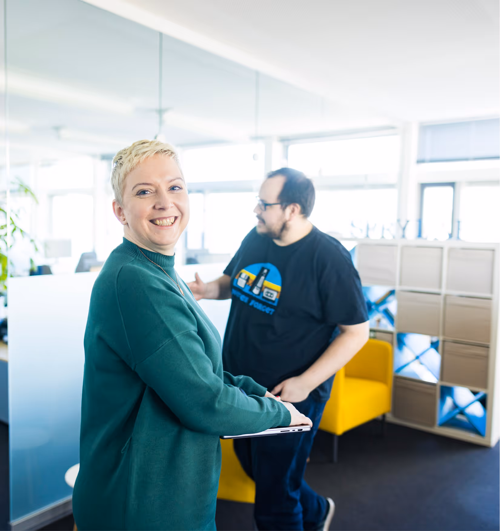Frau lächelt mit Laptop, im Hintergrund spricht ein Mann in einem modernen Büro.