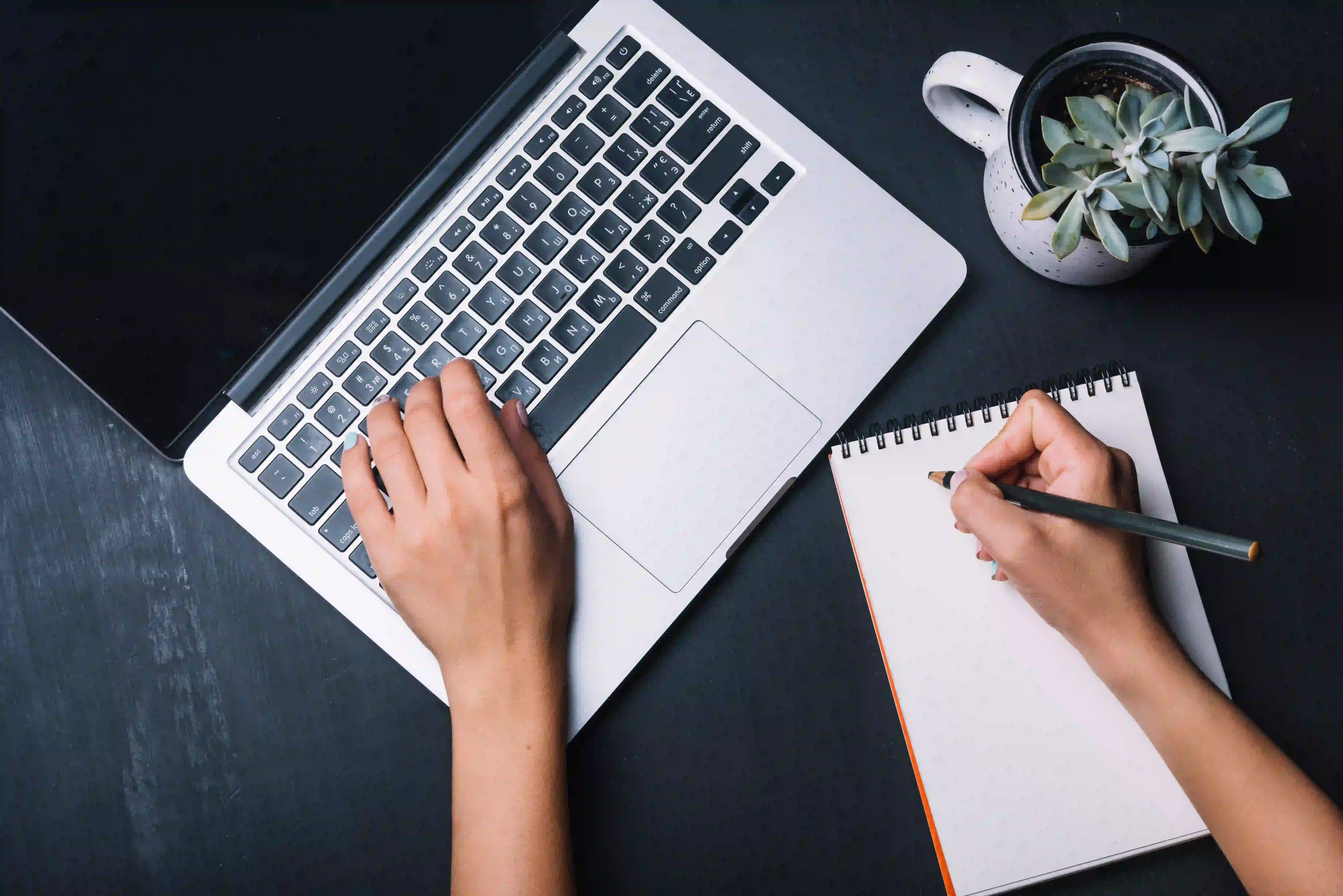 A person typing on a laptop and writing in a notebook, illustrating the dedicated scriptwriting process offered by a full explainer video company.