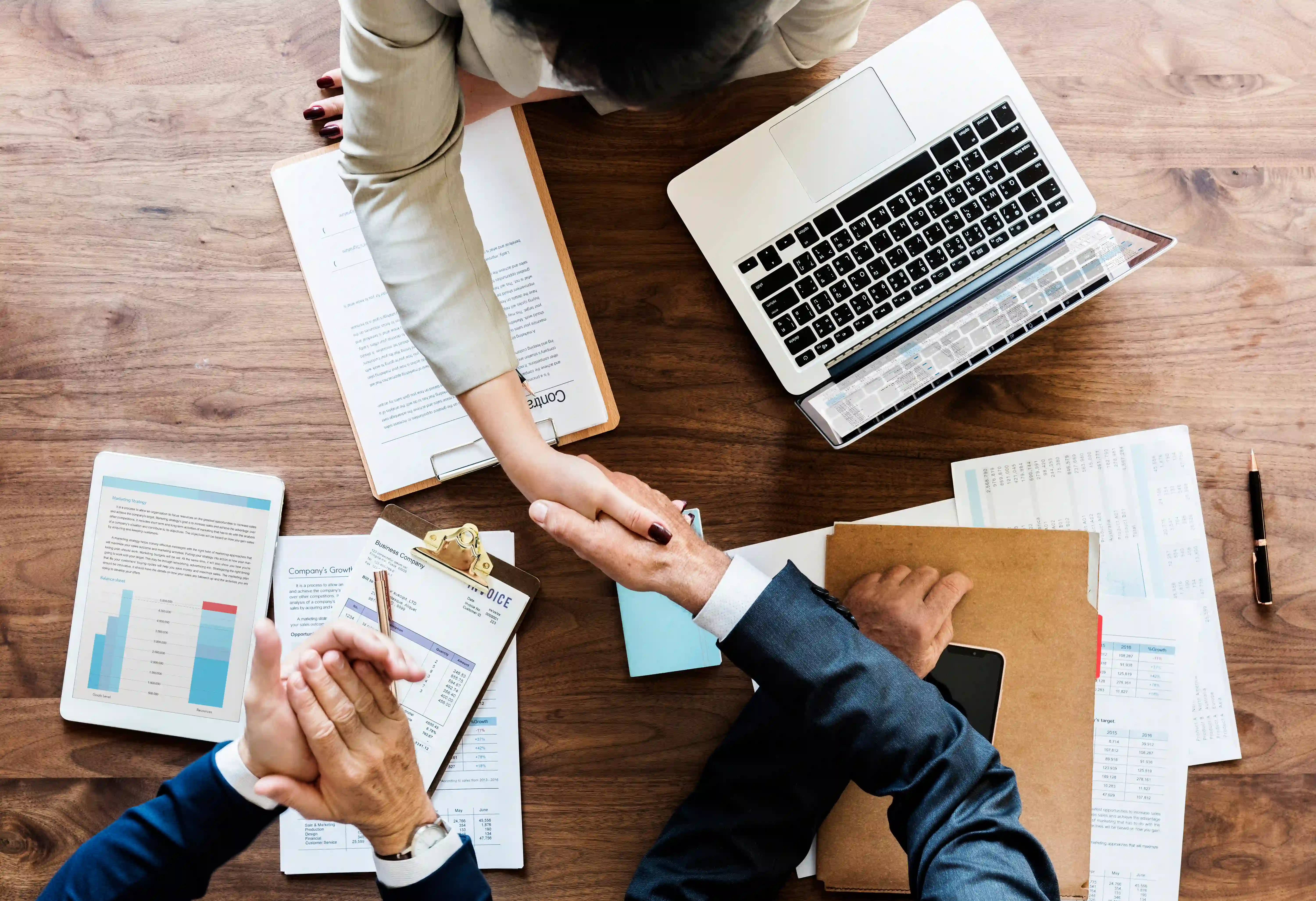 A top-down view of two professionals shaking hands across a desk with a laptop, representing the partnership between HR and marketing teams