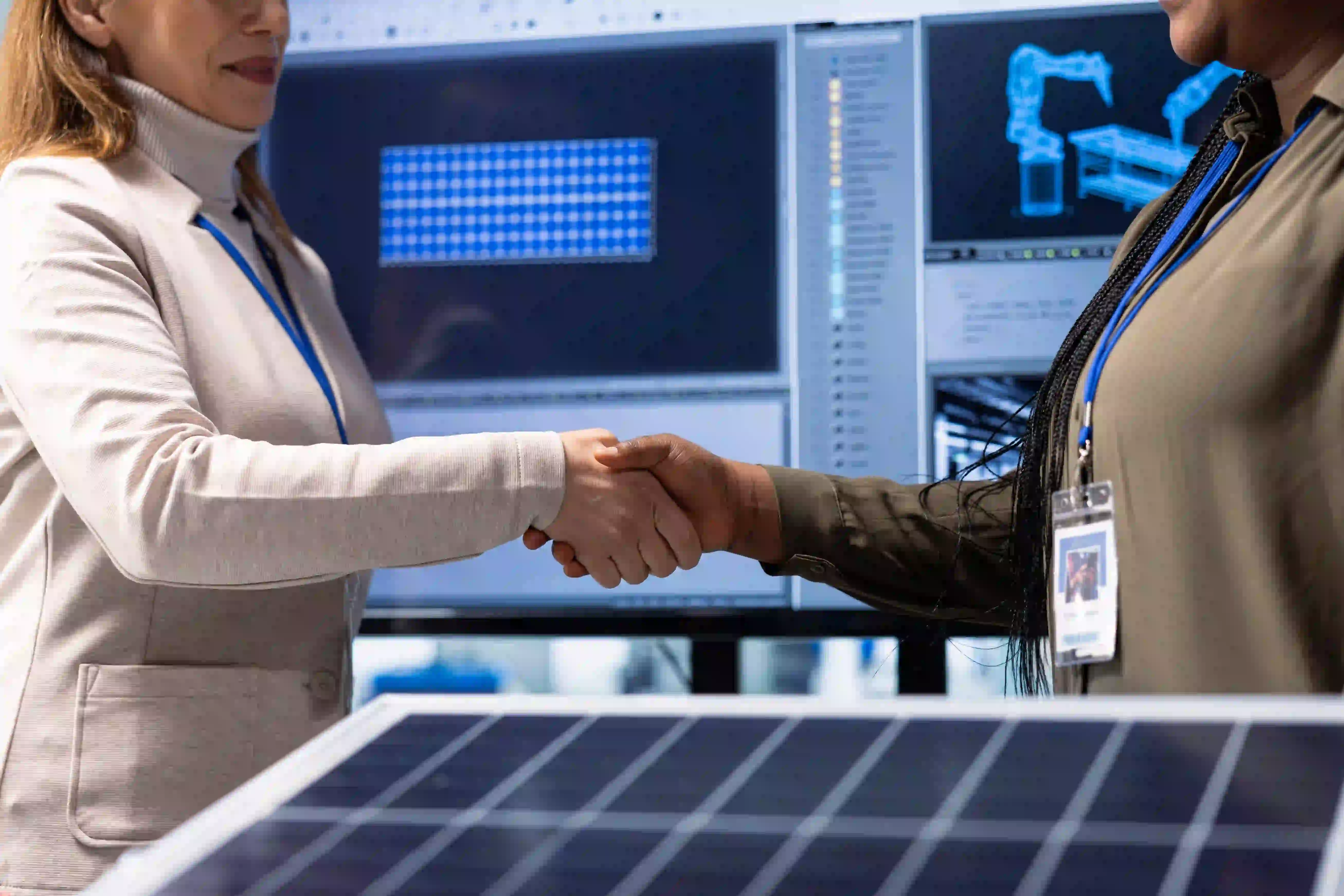 Two corporate professionals shaking hands in front of industrial screens, representing the ongoing alignment between sales and marketing teams as the business changes