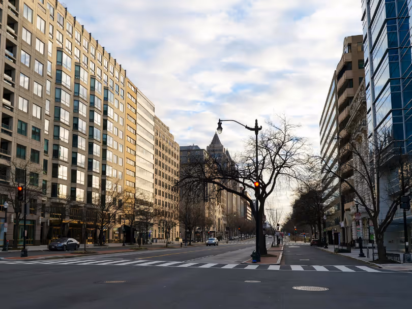 View of a city street lined with tall office buildings under a cloudy sky with leafless trees.
