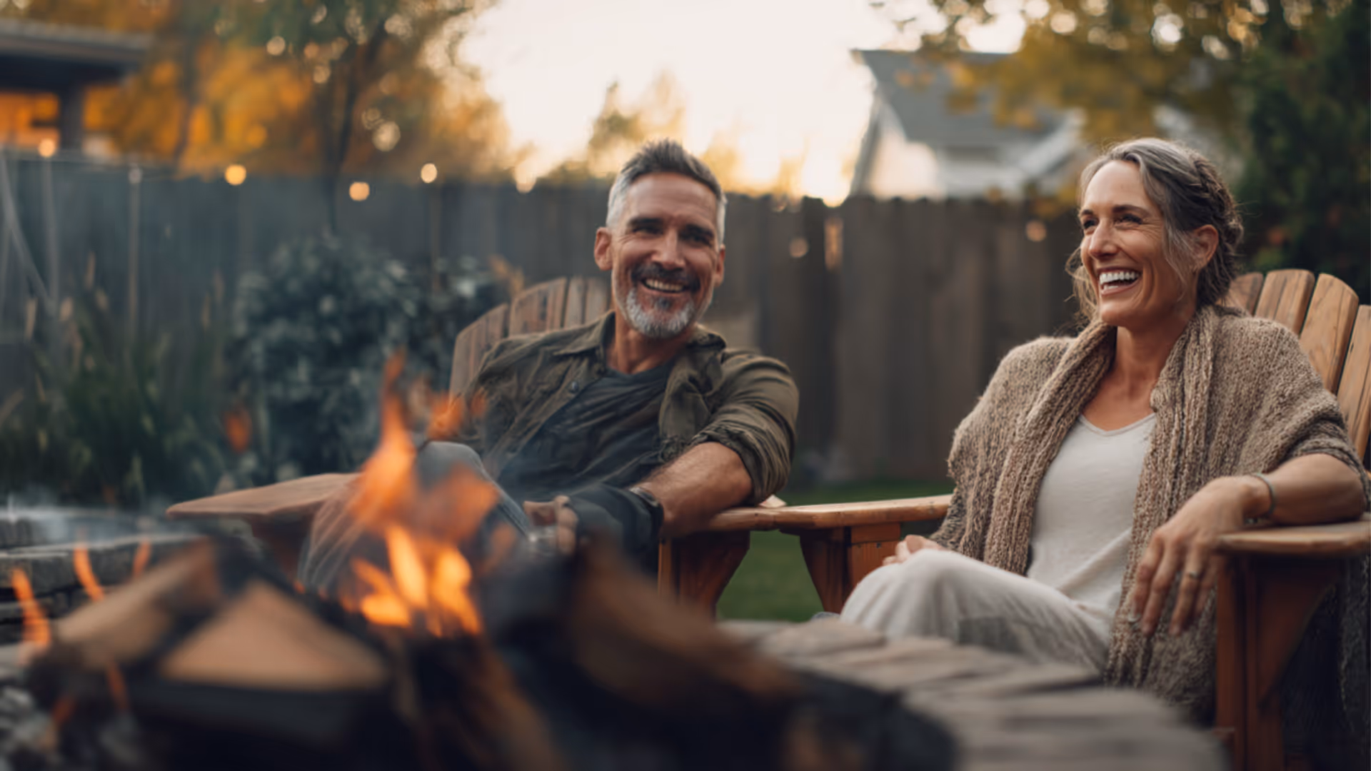 Smiling middle-aged man and woman sitting on wooden chairs by a fire pit outdoors in a backyard.