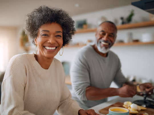 Smiling couple cooking together in a bright kitchen with food and a mug on the counter.