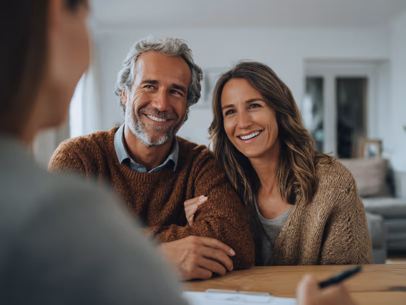Smiling middle-aged couple sitting at a table, talking with an advisor in a cozy home setting.